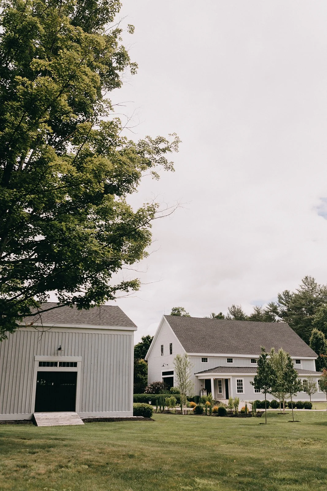 A large white farm-style house with a peaked roof, surrounded by a well-manicured lawn and small trees, with a gray outbuilding nearby. The sky is overcast.