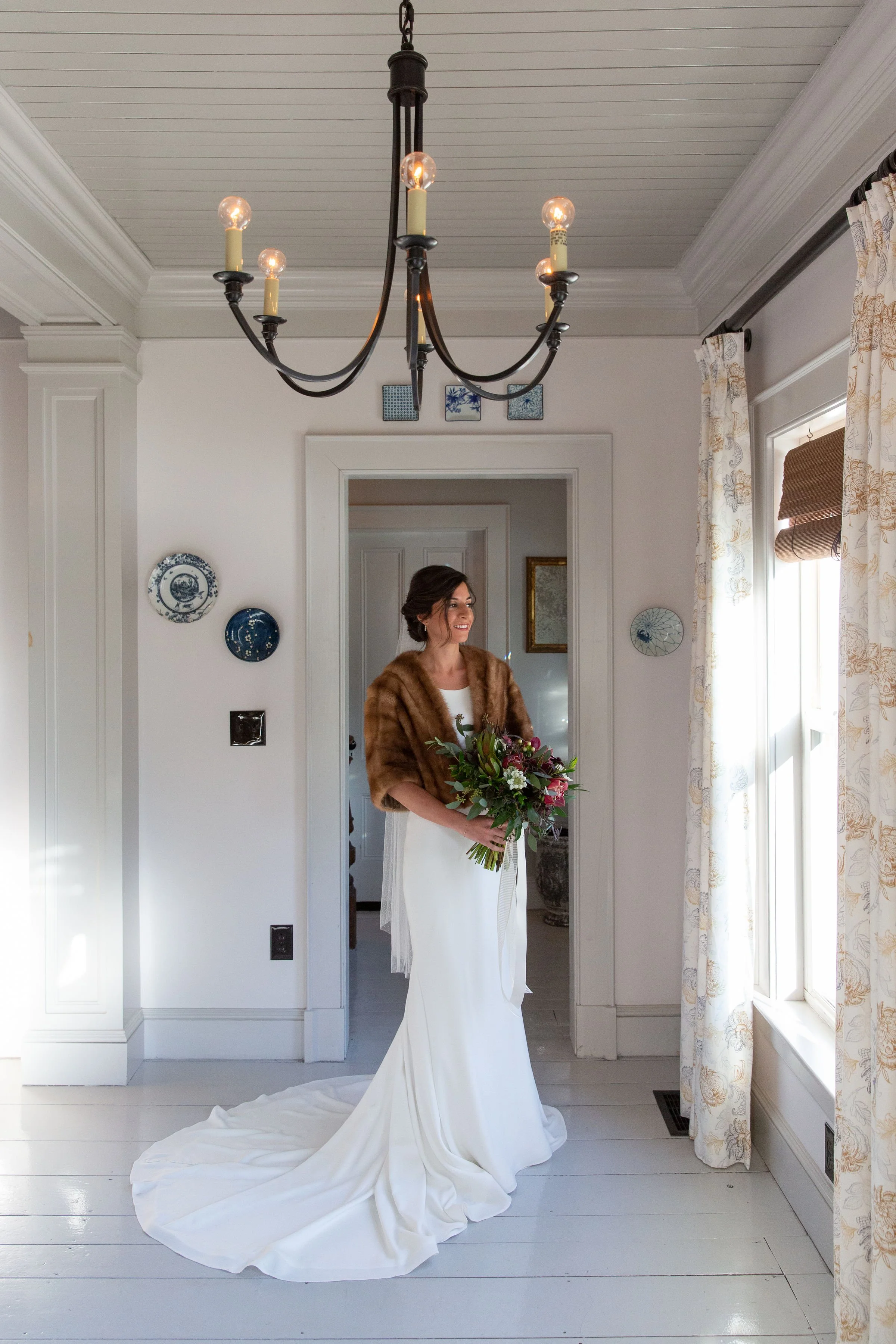 A woman in a wedding dress holding a bouquet of flowers standing in a bright room with white walls, decorated with blue and white plates, and curtains on the windows.