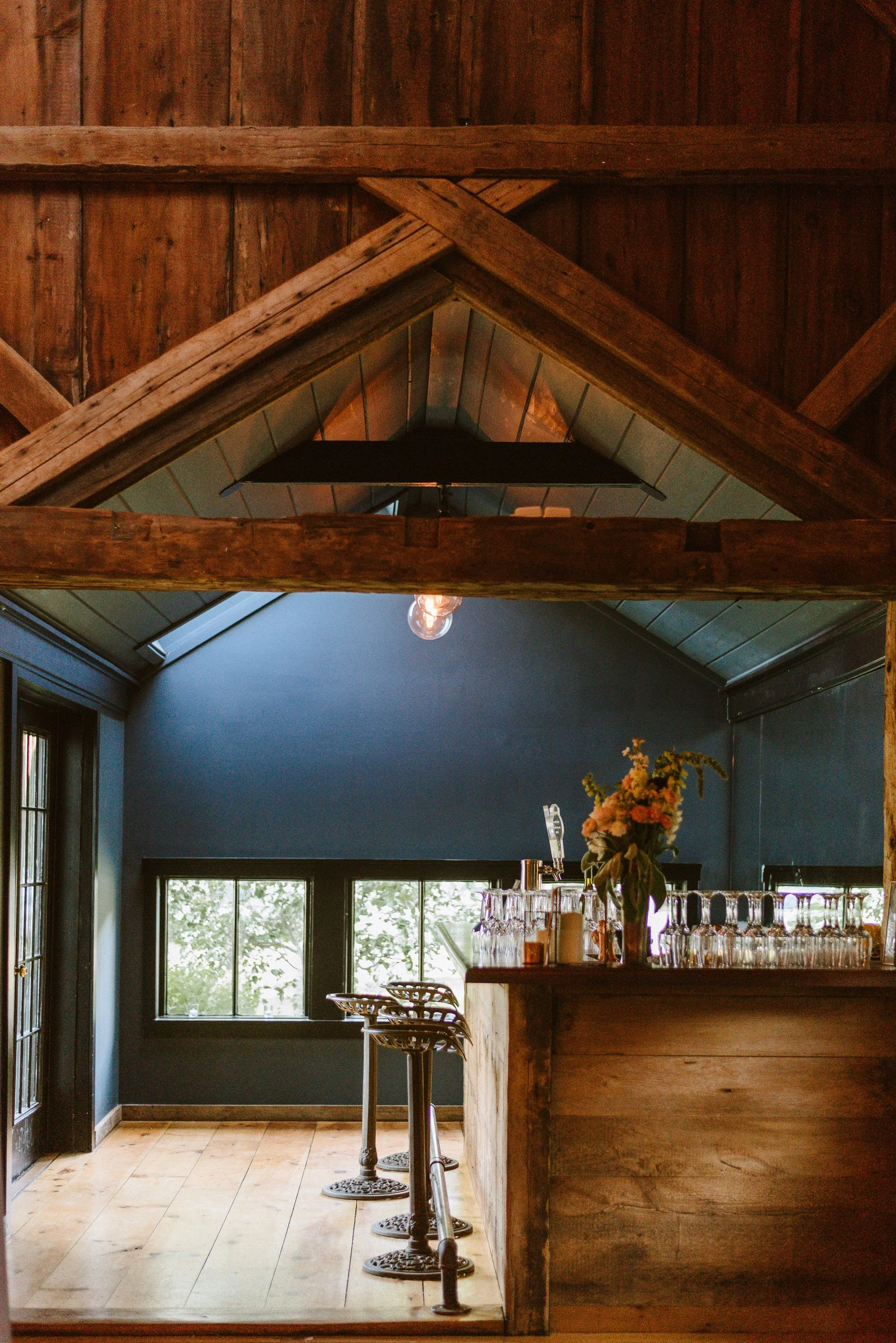 Interior of a rustic bar with a wooden counter, barstools, a window with daylight, and a flower bouquet on the bar.