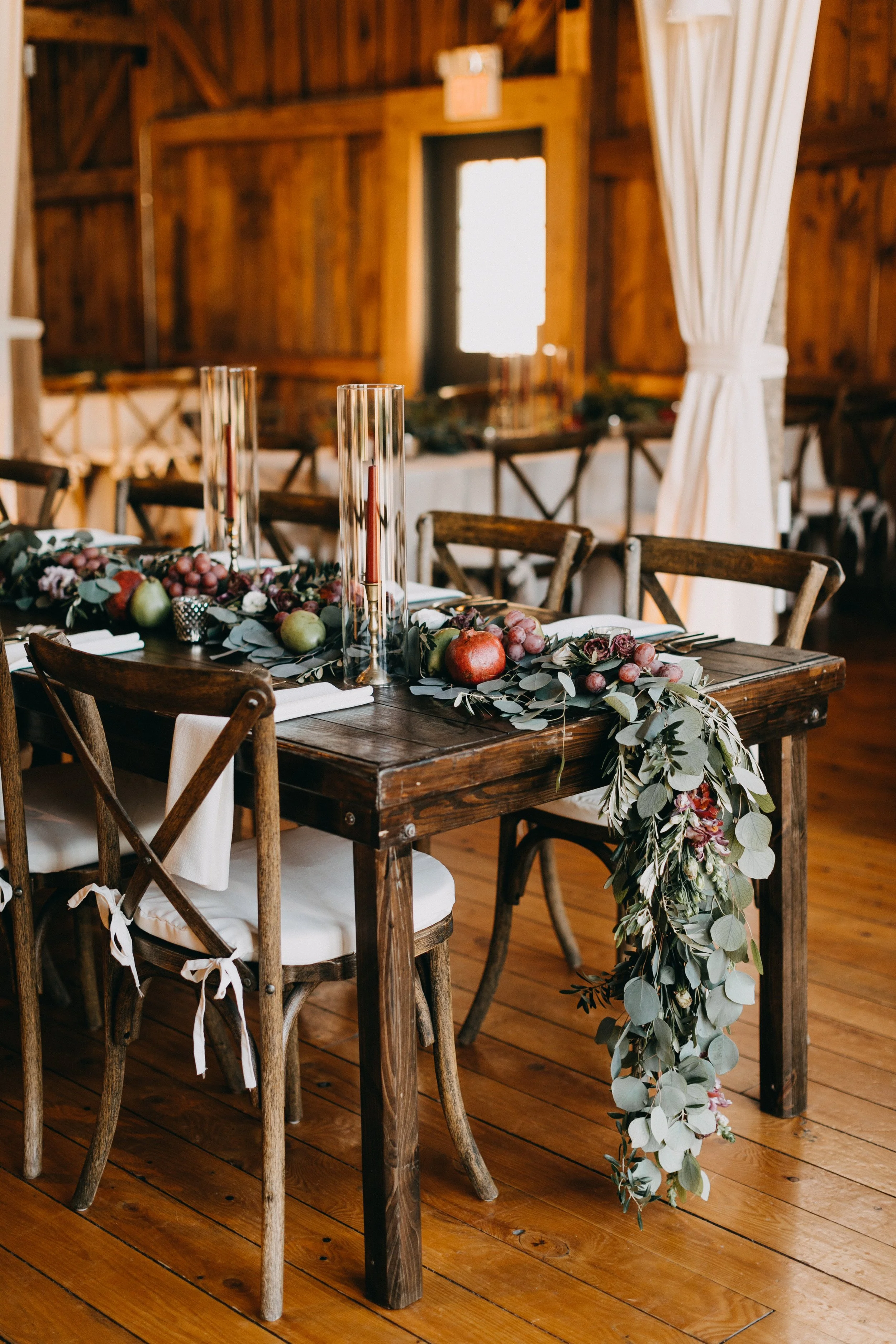 A rustic wooden table decorated with a greenery and fruit garland, tall glass candle holders, and white napkins, set inside a barn-like room with wooden walls and floors, and white curtains.