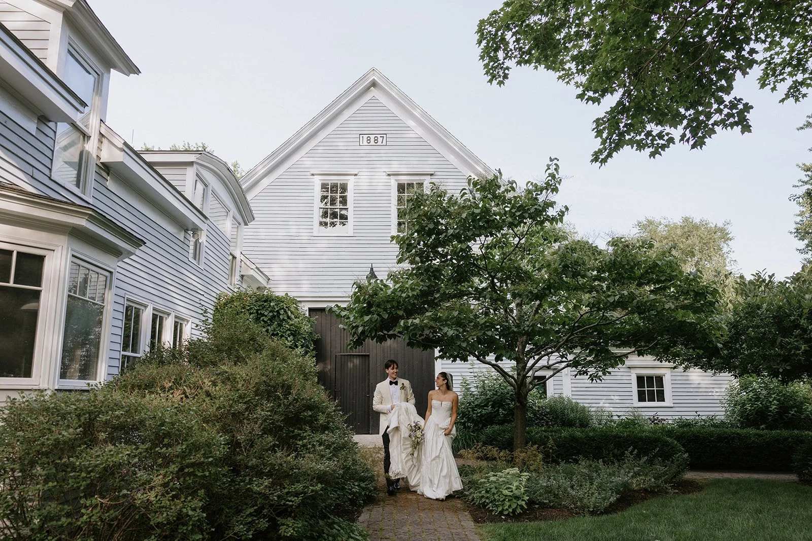 A bride and groom in wedding attire walking together outside a large white house with garden, trees, and shrubs.