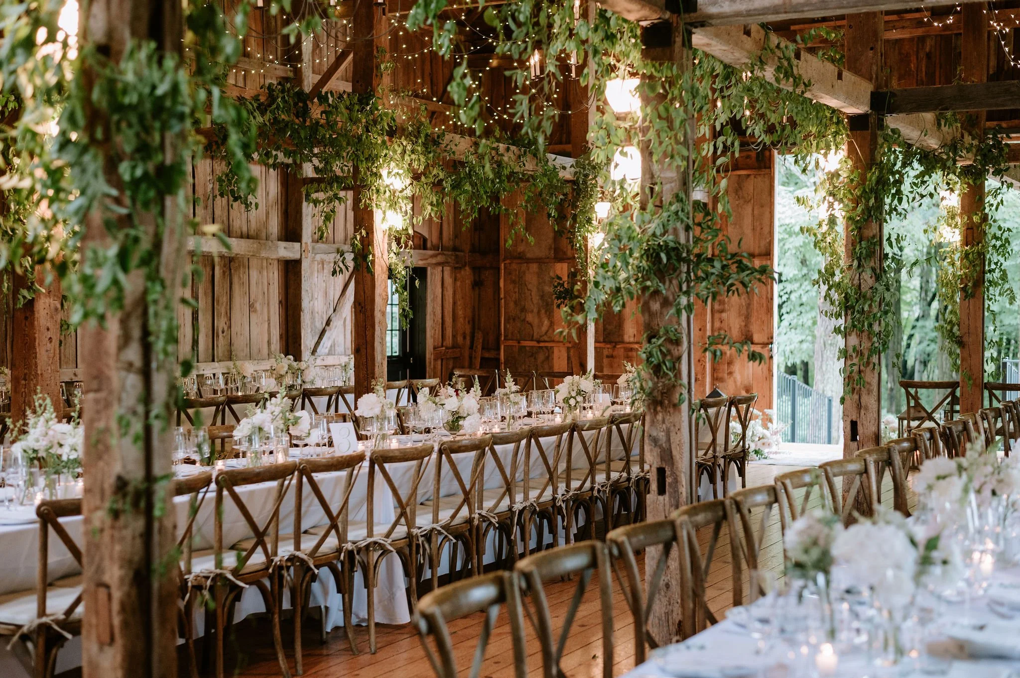 Wedding reception setup with long banquet tables, decorated with white flowers and glassware, inside a rustic barn with wooden walls and green foliage hanging from the ceiling.