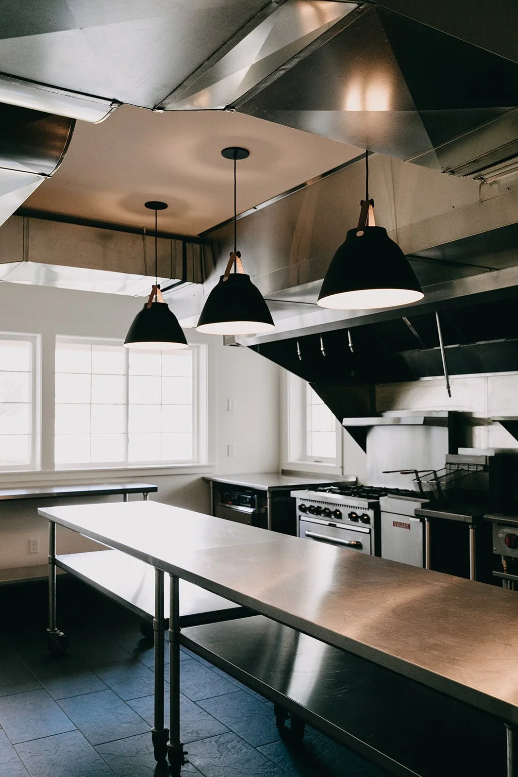 Modern commercial kitchen with stainless steel tables, black pendant lights, and industrial stove under a stainless steel range hood.