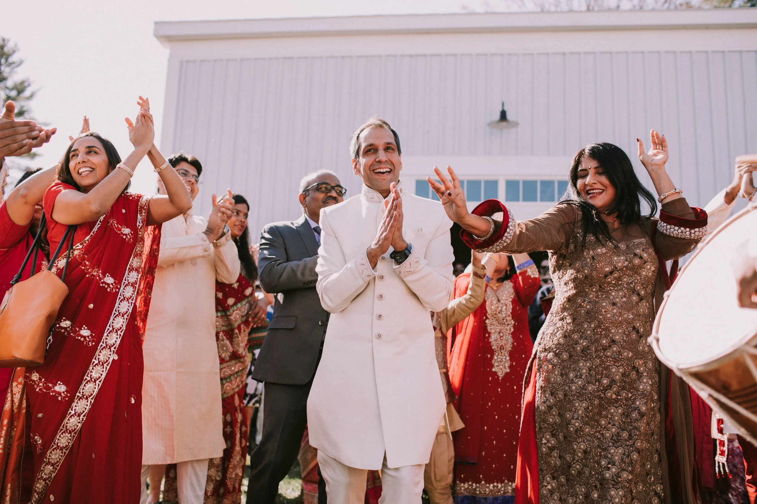 A group of people at a celebration, some wearing traditional Indian attire, with a man in a white Indian suit at the center, clapping and smiling, outdoors in front of a white building.