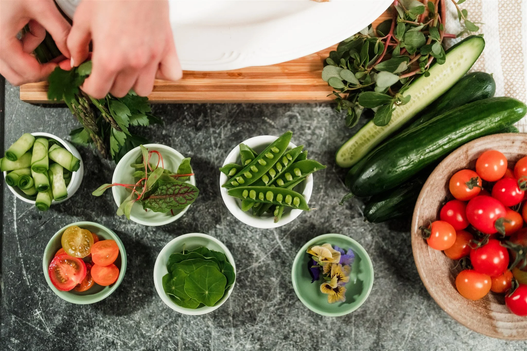 Overhead view of fresh vegetables and herbs on a gray marble surface, including cherry tomatoes, cucumbers, zucchinis, and various microgreens and edible flowers in small bowls.