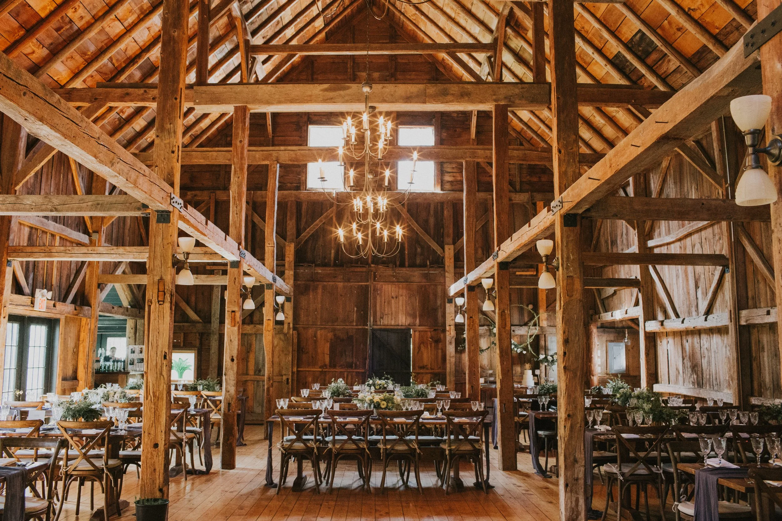 Interior of a rustic wooden barn decorated for an event with long tables and chairs, floral centerpieces, and a chandelier hanging from the high ceiling.