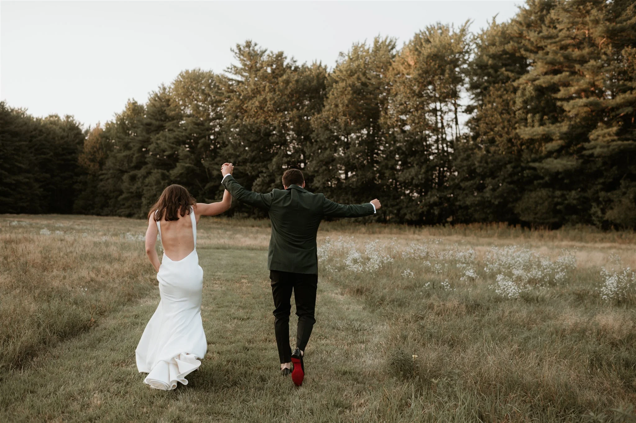 A couple dressed in wedding attire walking through a grassy field holding hands, with trees in the background during the evening.