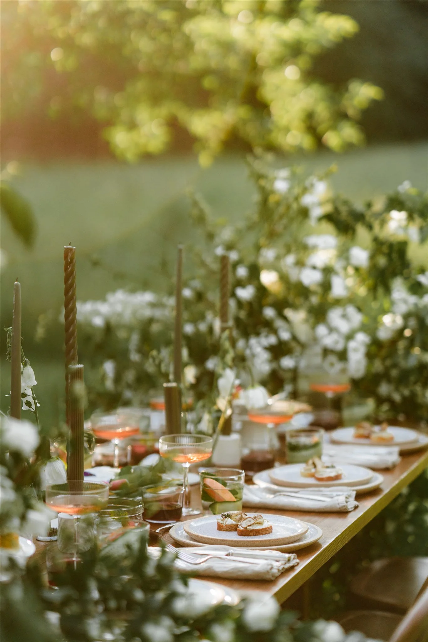 Outdoor dining table set with white plates, glasses, and silverware, decorated with flowers, candles, and greenery.