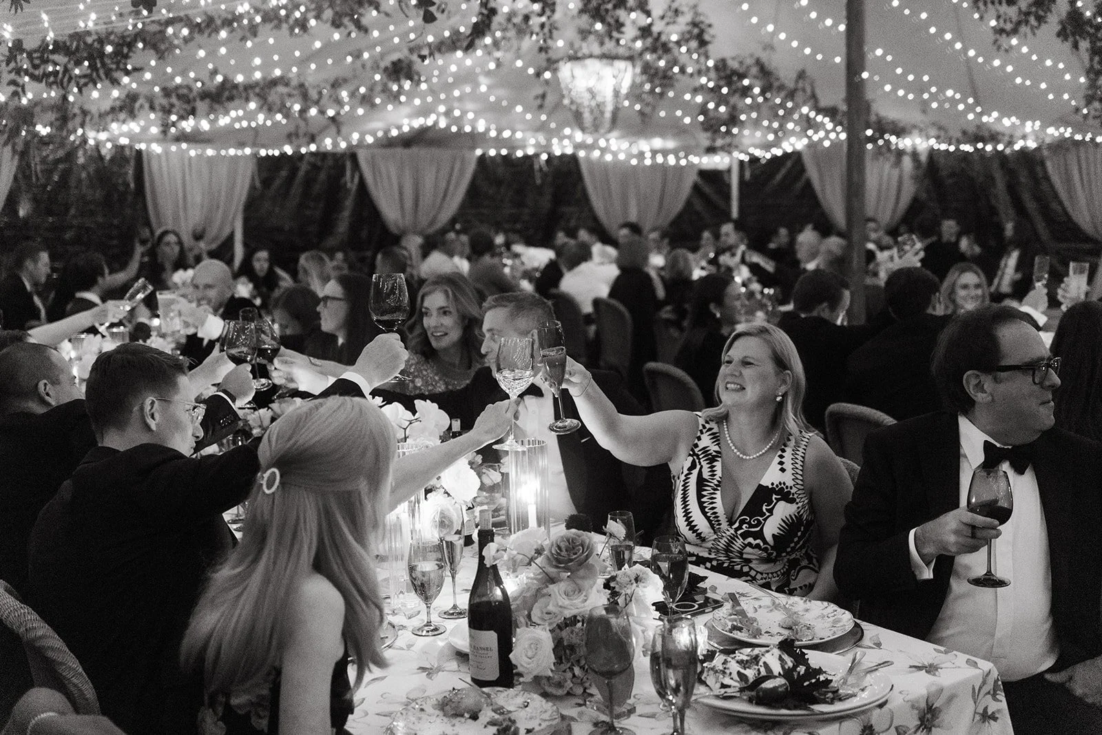 People at a formal celebration raising glasses in a toast, sitting around a decorated table under string lights and draped curtains.
