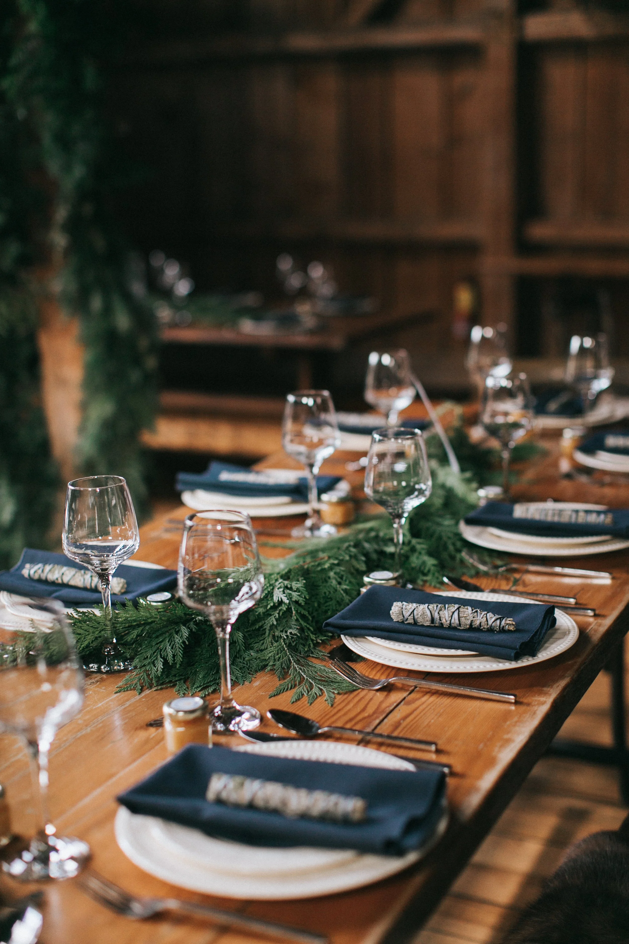 Decorated wooden dining table set with white plates, navy napkins, silverware, and empty wine glasses, adorned with a green holiday-themed garland for a festive occasion.