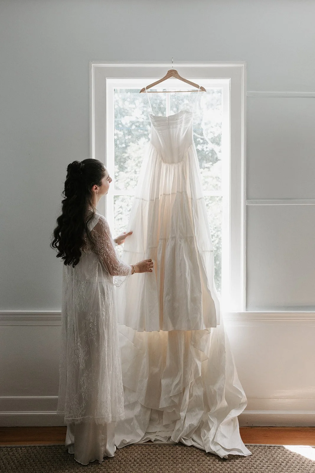 A woman in a lace dress looks at a hanging wedding dress in front of a window.