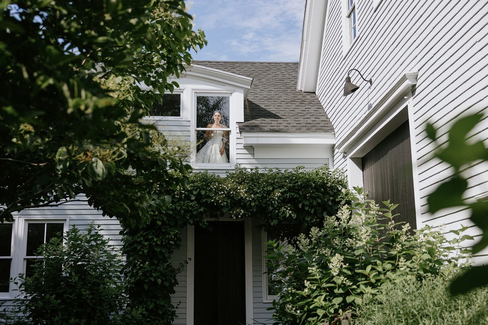 A woman in a wedding dress looking out a second-story window of a white house, surrounded by greenery and plants.