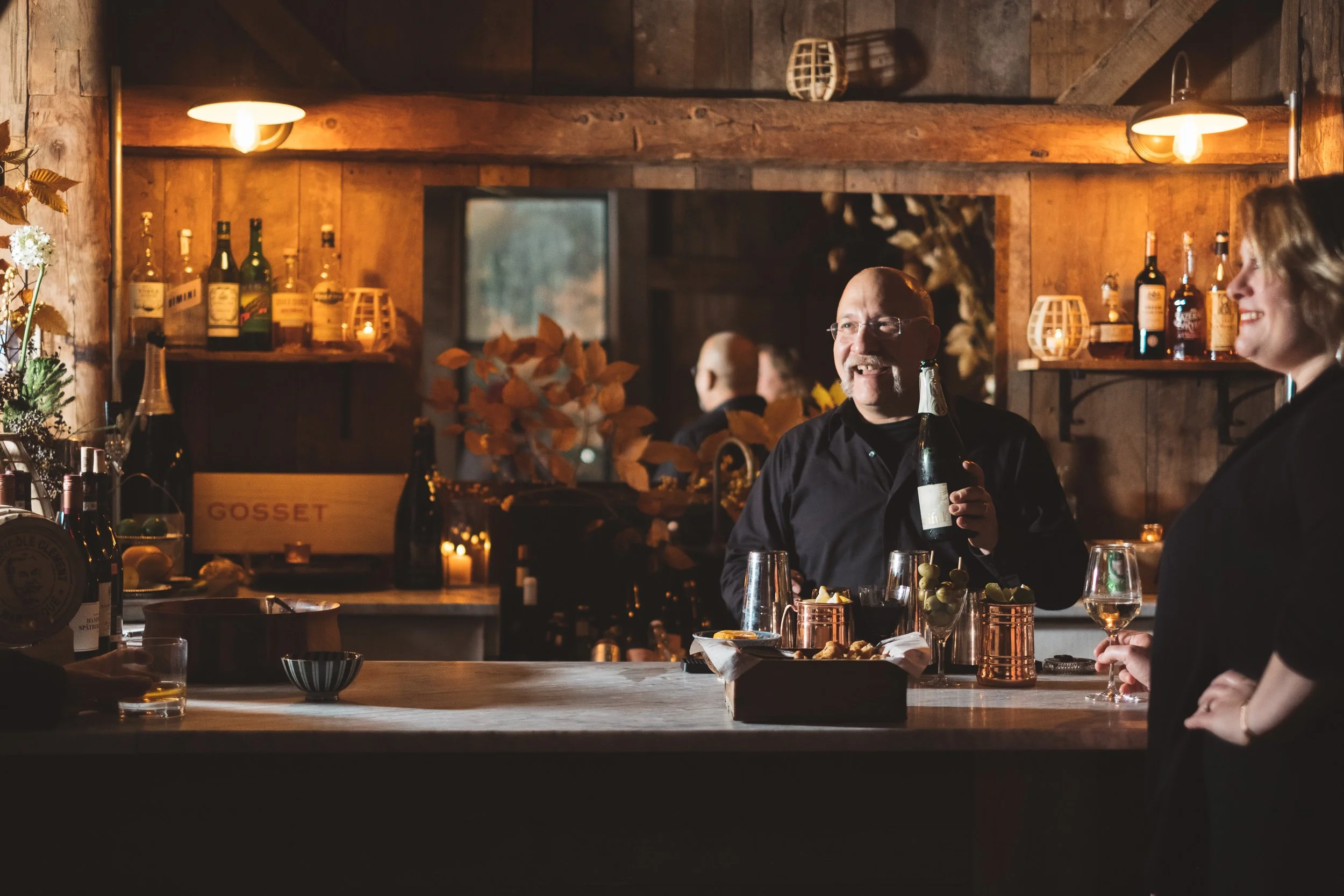 A man holding a bottle of champagne and smiling at a woman in a black dress in a warmly lit wooden bar with bottles, glasses, and candles.