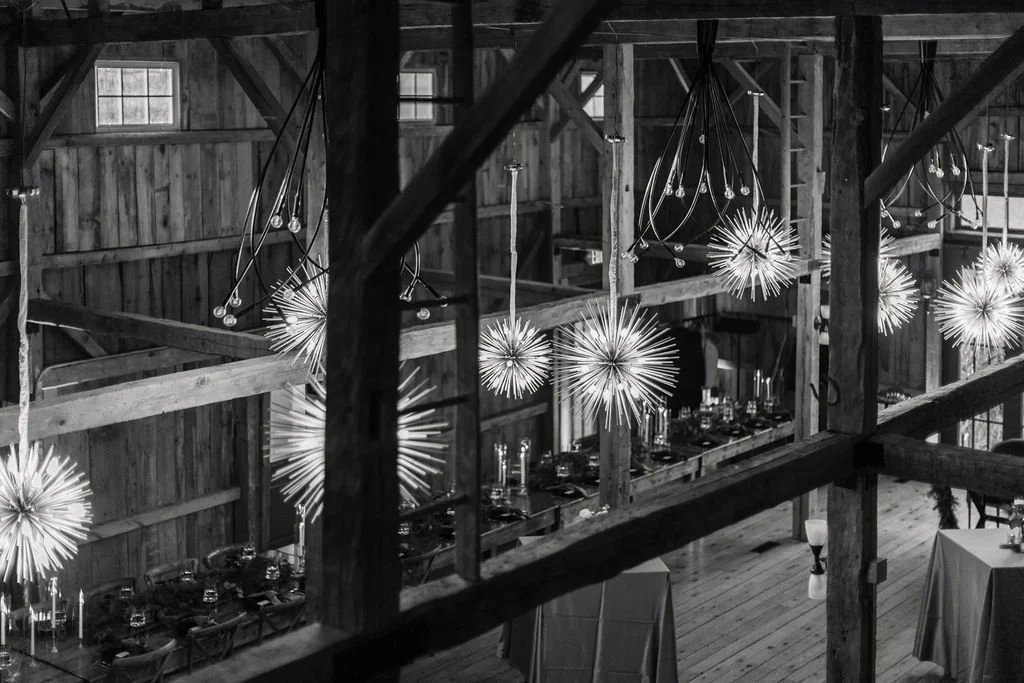 Interior of a rustic barn decorated with modern hanging light fixtures, including spherical starburst designs, over a long dining table set for an event.