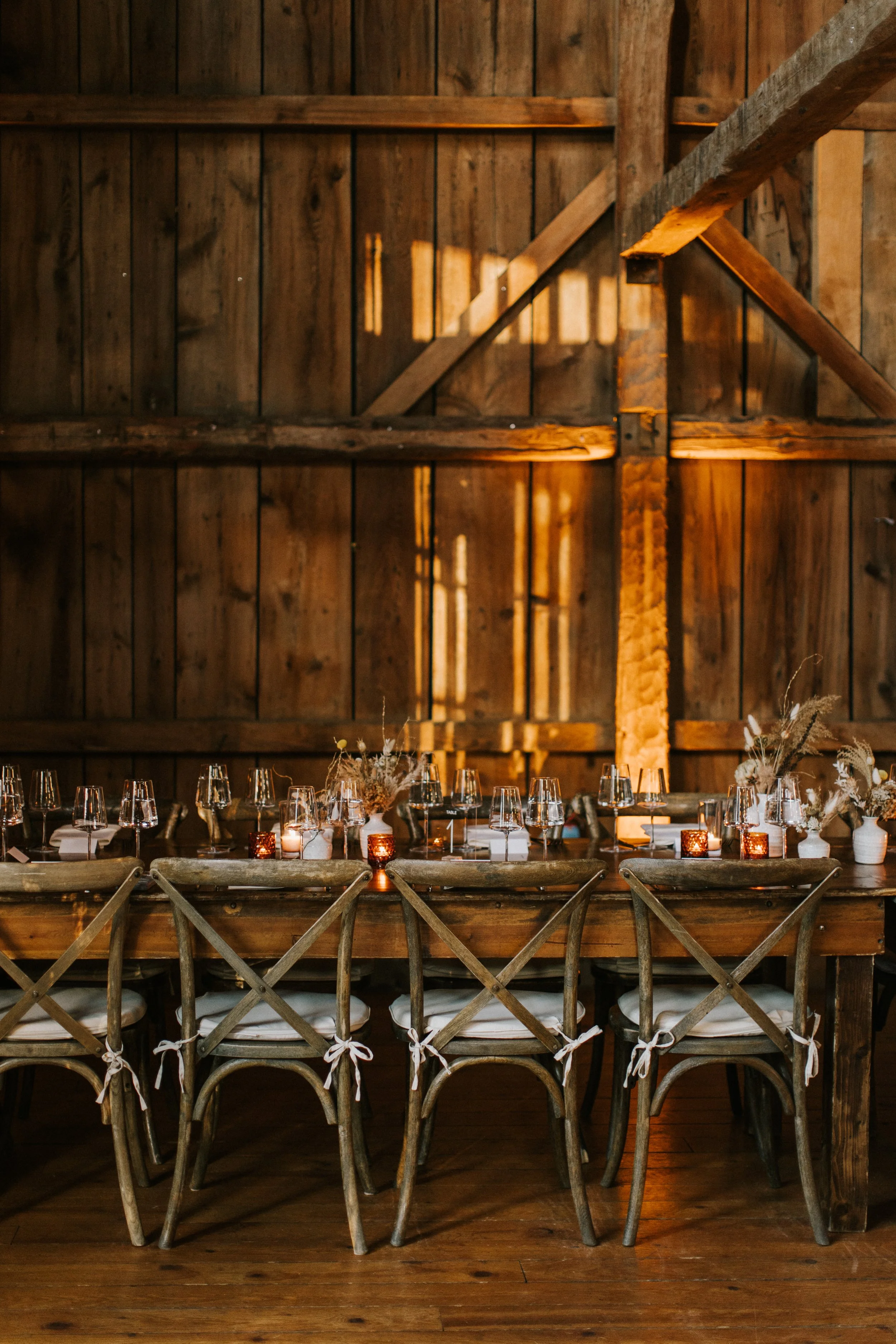 An elegantly set dining table inside a rustic barn with wooden walls, decorated with glasses, candles, and dried floral arrangements.