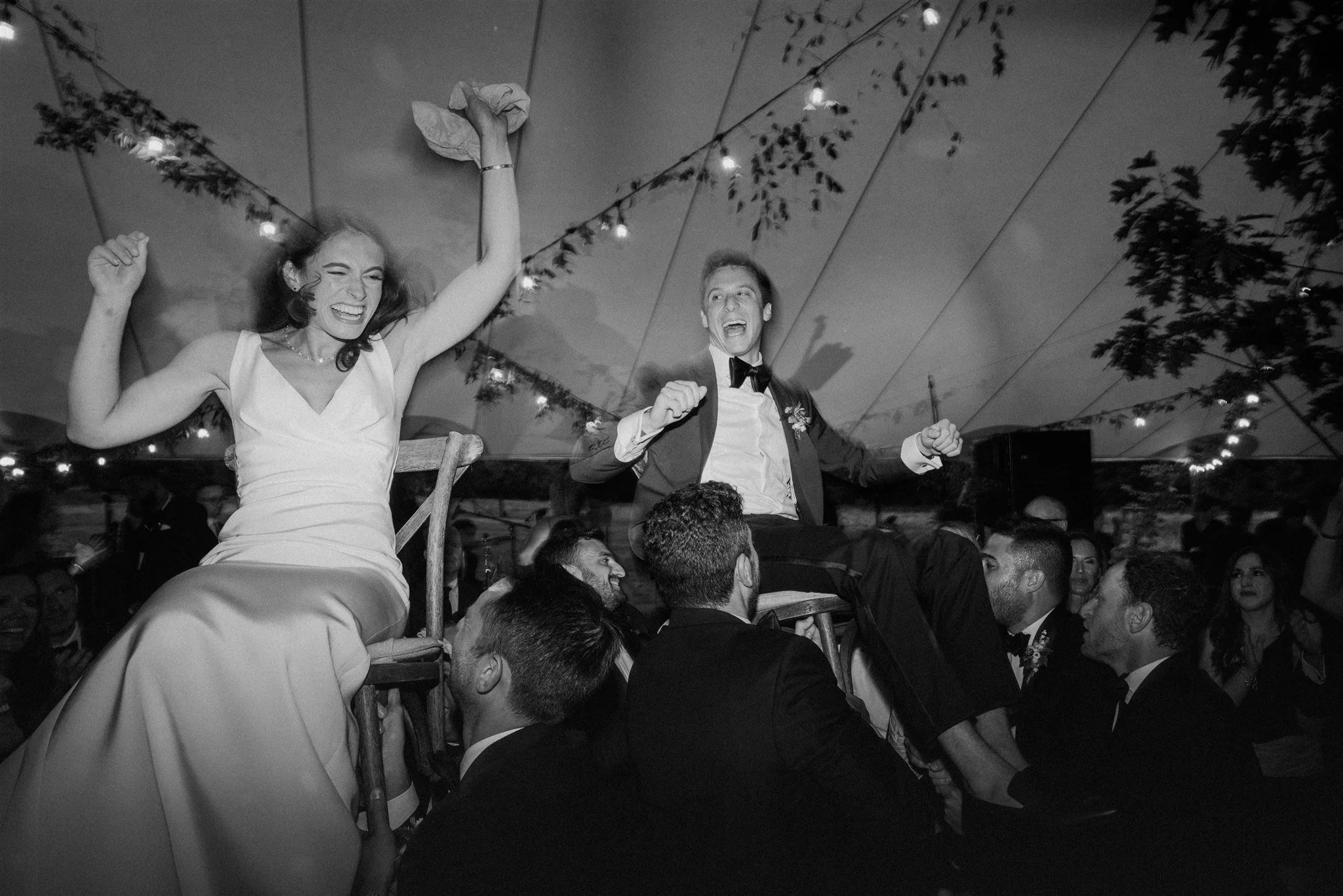 Black and white photo of a wedding celebration, with a woman in a wedding dress and a man in a tuxedo being lifted on chairs by a group of men, surrounded by guests, under string lights and a tent canopy.