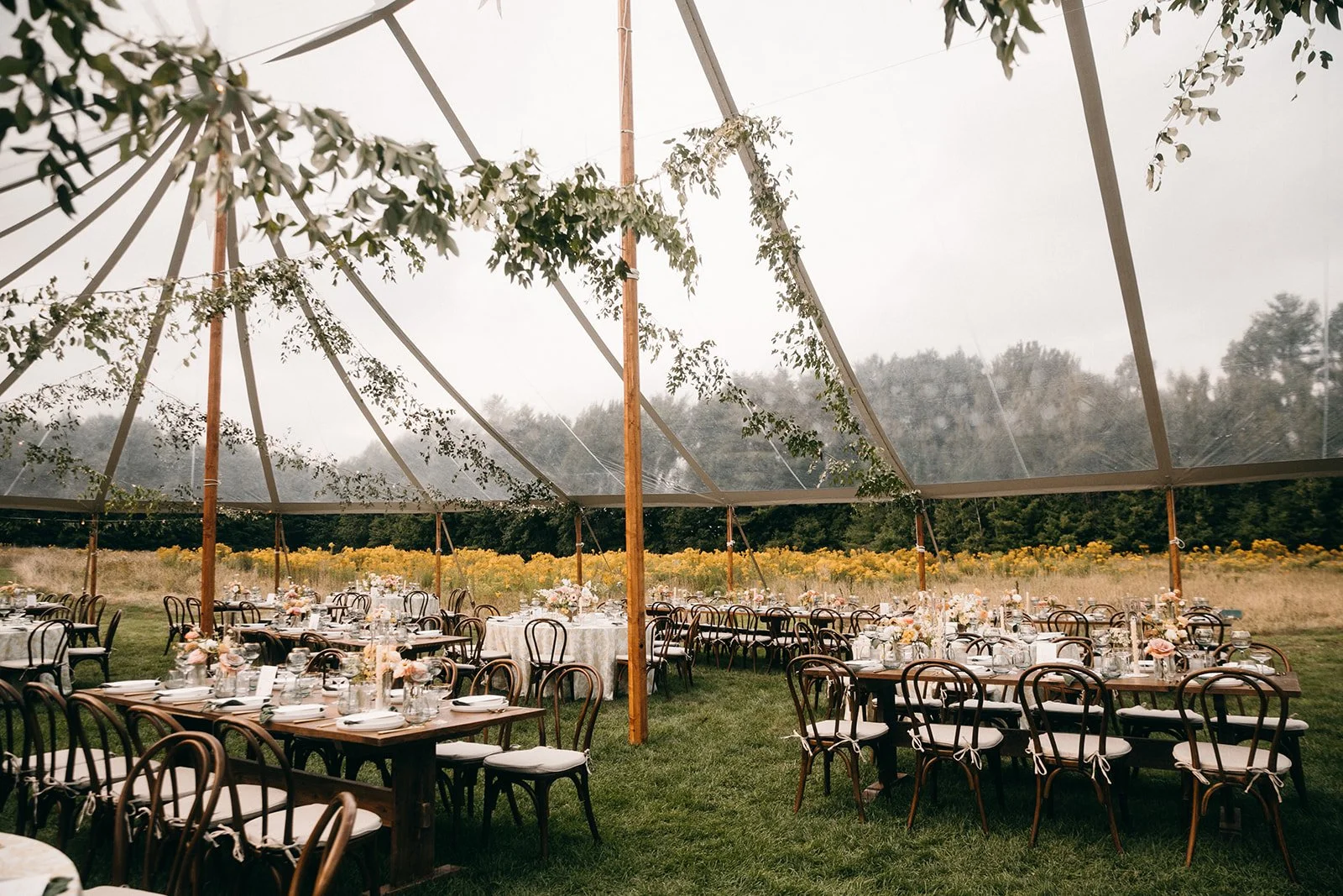 Outdoor wedding reception setup with decorated tables and chairs under a large tent with hanging greenery, on a grassy field during daytime.