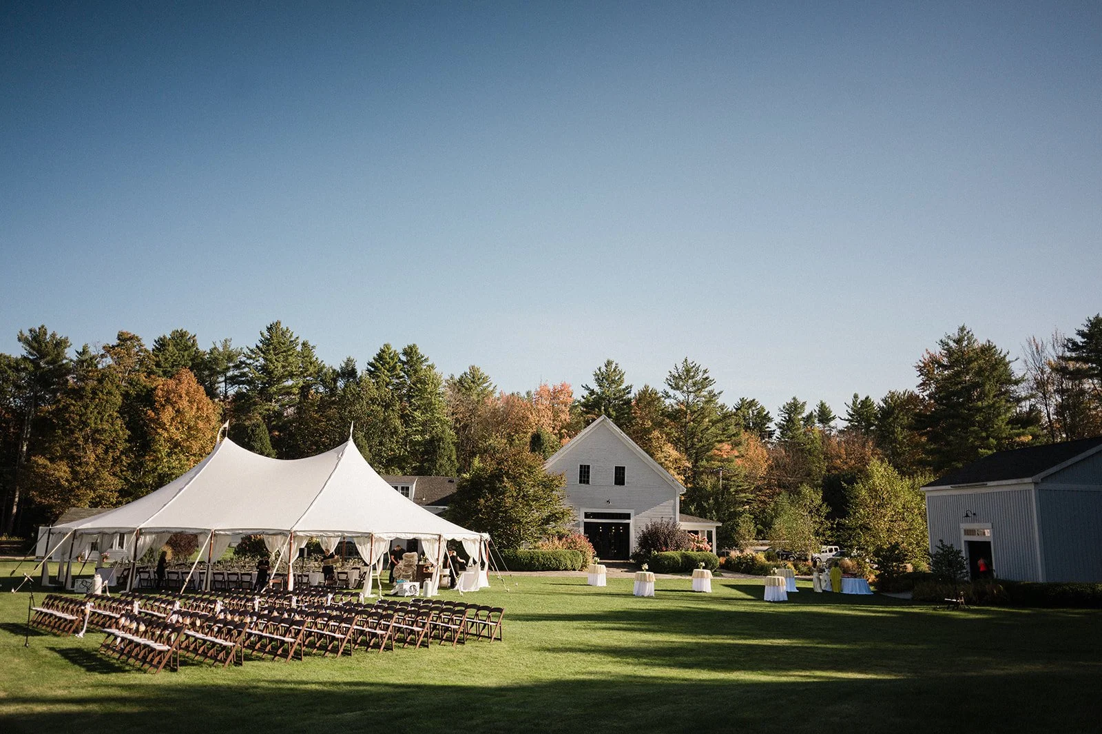 An outdoor wedding venue with a large white tent set up on a grass lawn, surrounded by trees and a white building in the background.