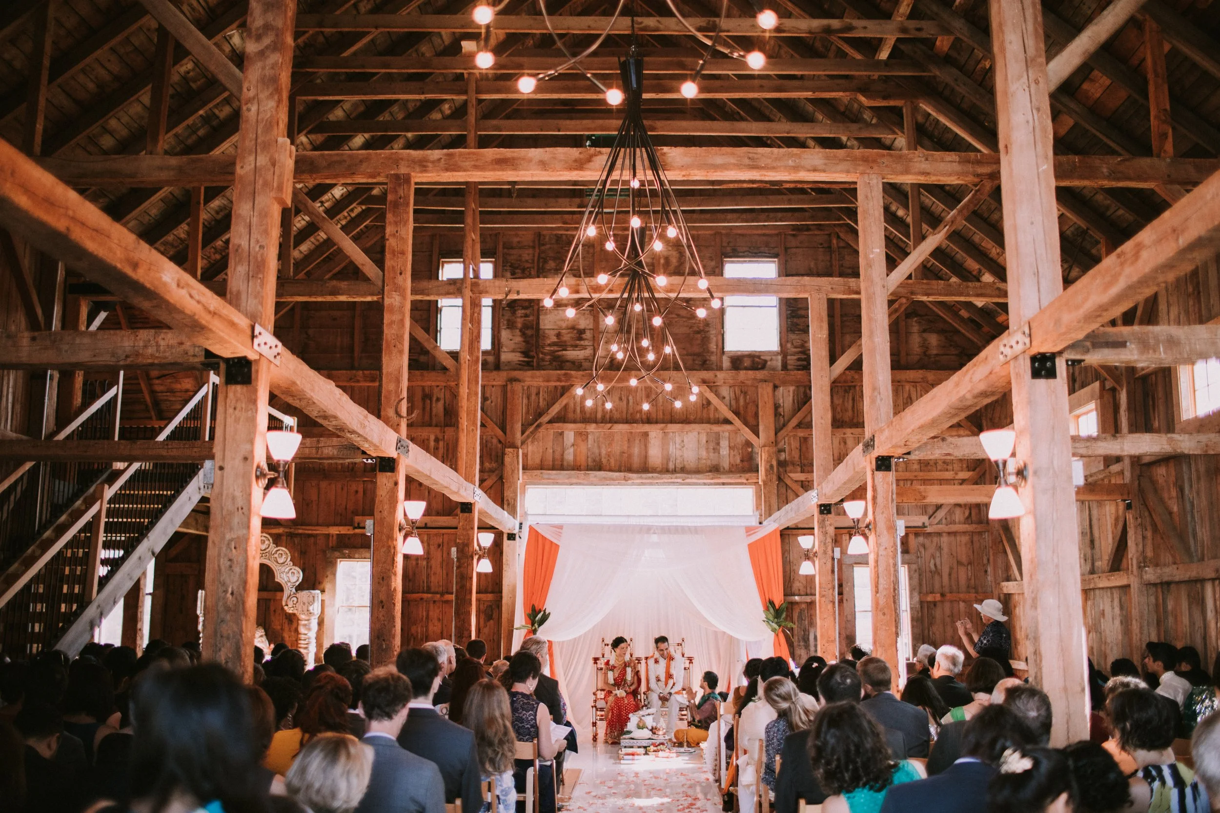 A wedding ceremony in a rustic barn with wooden beams, string lights, and a couple seated at the front under a white and orange draped backdrop.