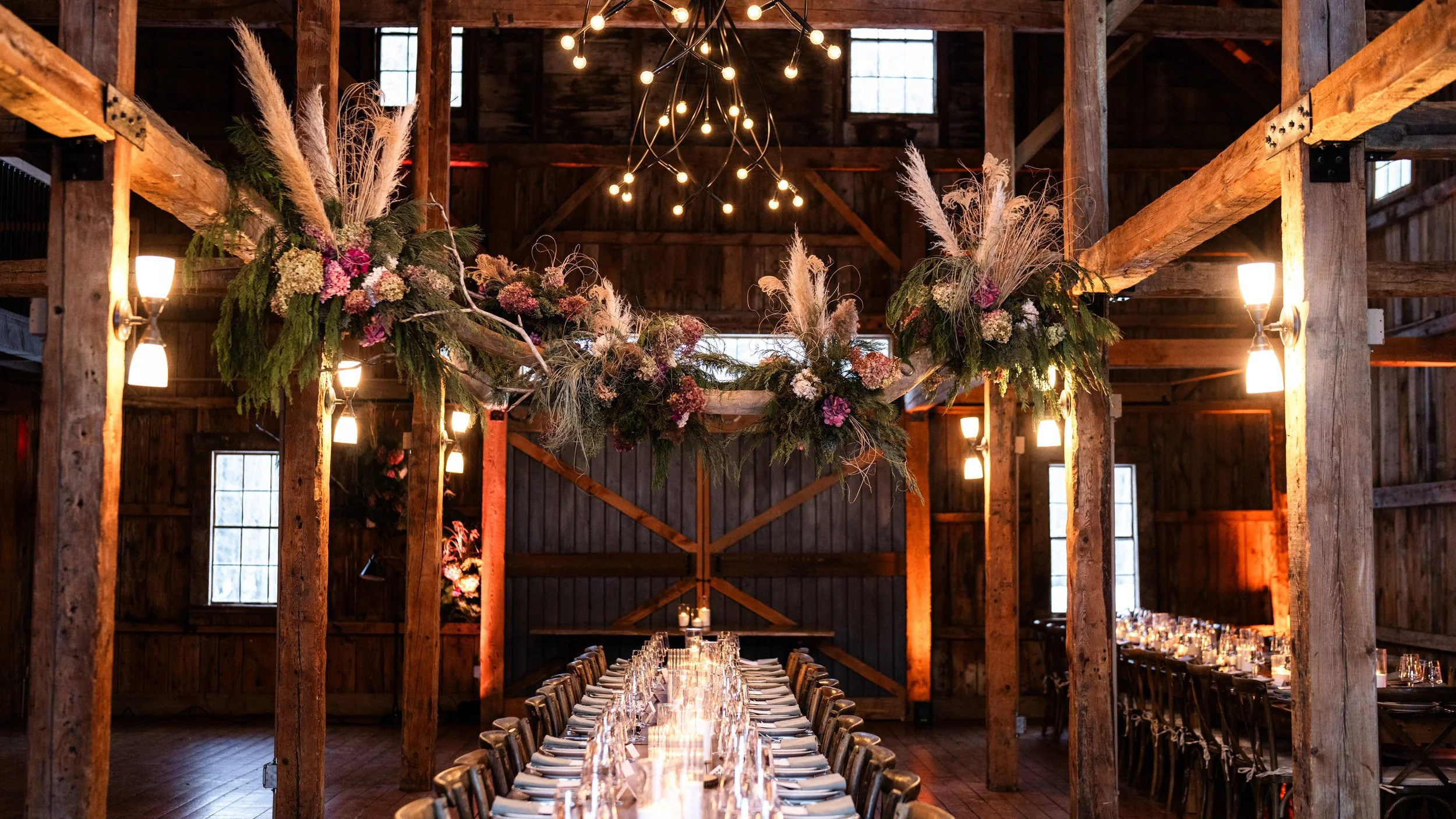 Long dining table in a rustic barn wedding venue decorated with floral arrangements and candlelight
