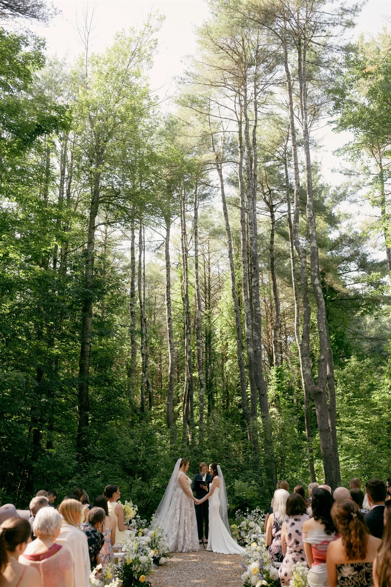 A wedding ceremony taking place outdoors in a forest with tall trees, with two brides in white wedding dresses holding hands, standing in front of an officiant, surrounded by seated guests.