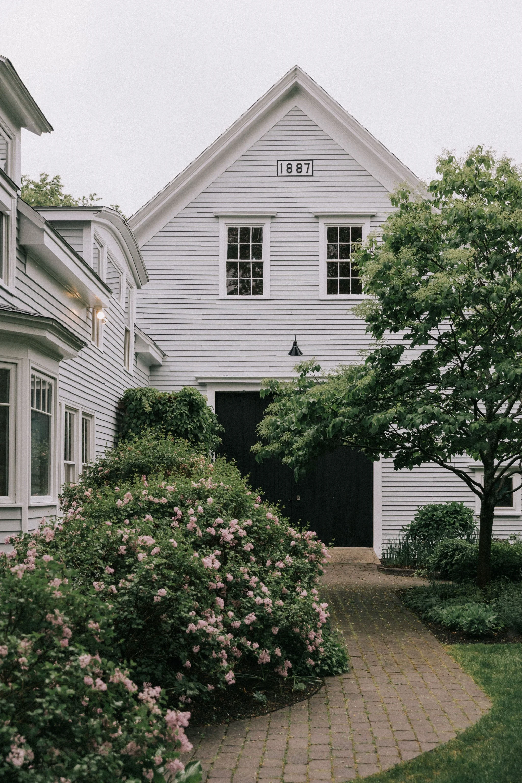 Front view of a white farmhouse with black doors, a gabled roof, and two windows on the upper floor, with the date 1887 displayed. Green bushes and trees line a stone pathway leading to the entrance.