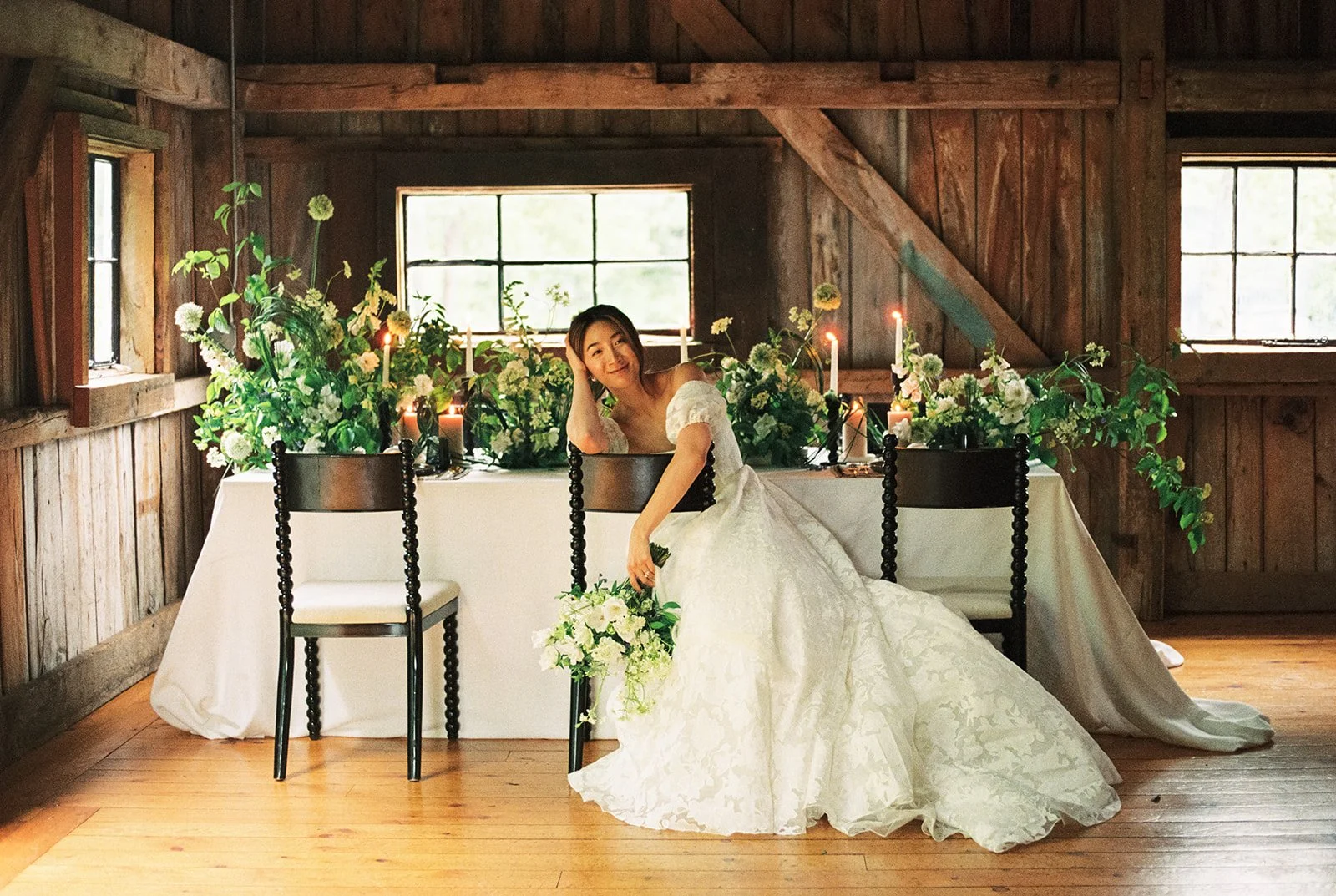 A woman in a white wedding dress sitting and leaning on a chair at a decorated table with green and white floral arrangements inside a wooden room with windows.