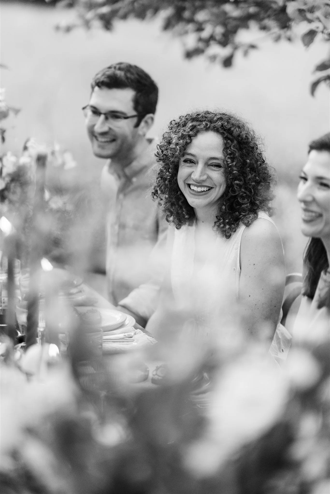 Black and white photo of four people enjoying a meal outdoors, smiling and laughing, with a blurred background of trees and foliage.