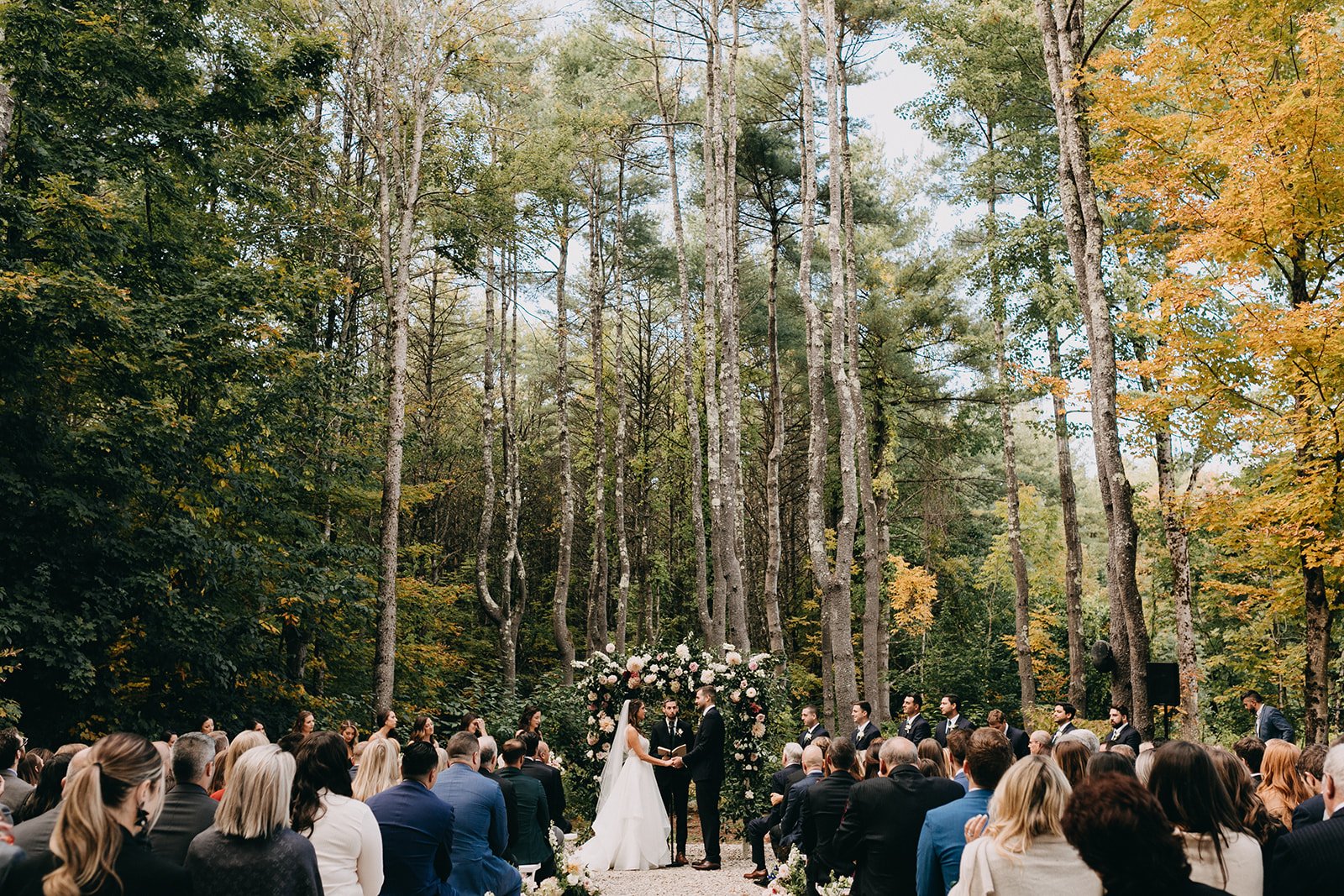 Wedding ceremony outdoors in a forest, with the bride and groom holding hands in front of guests and a floral arch.