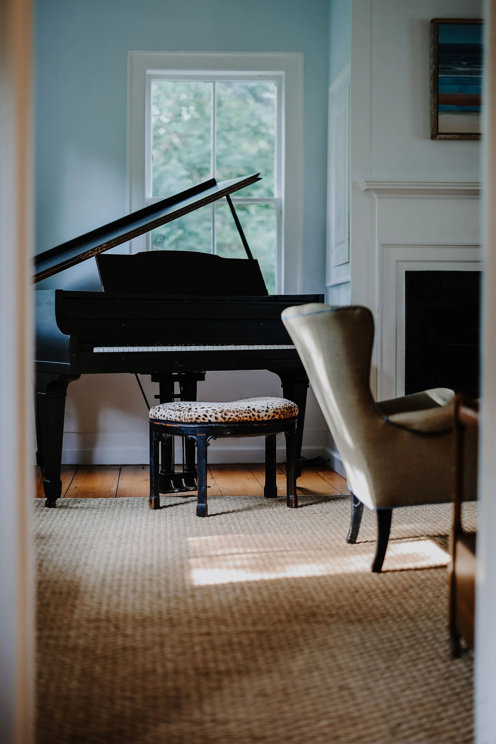 A black grand piano in a room with wooden floor, white walls, a window, and a chair with a leopard print cushion.