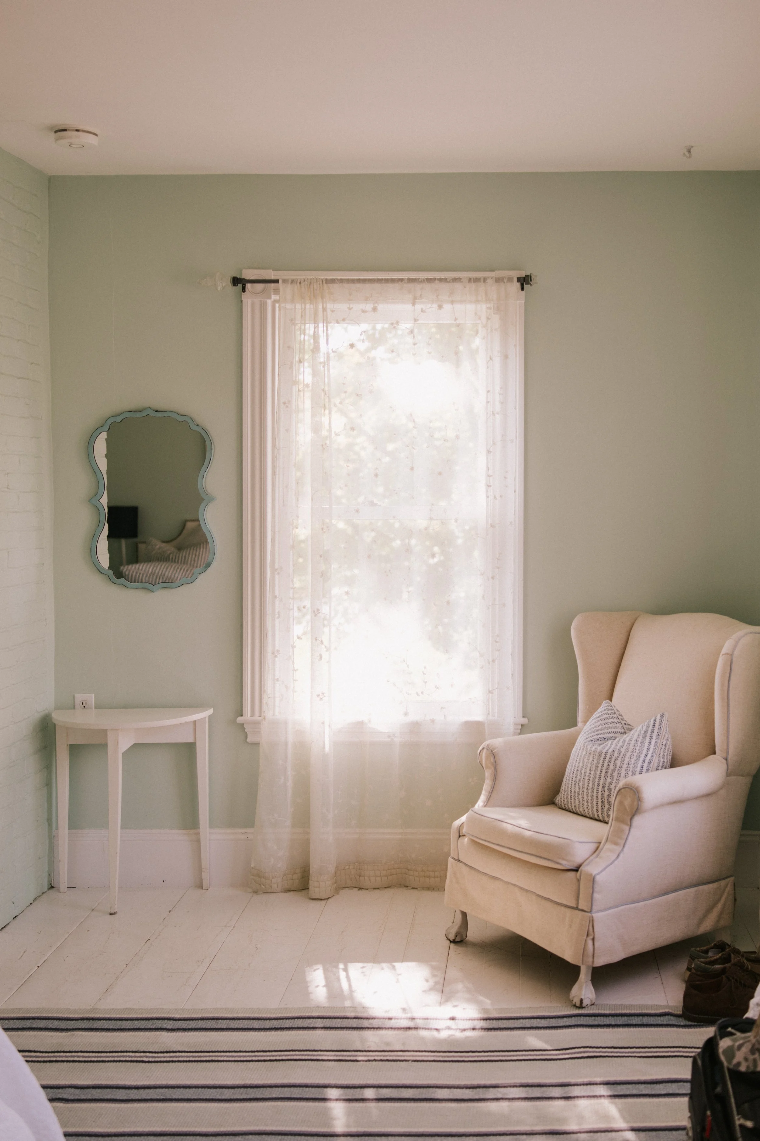 A cozy corner of a room with a large window covered by sheer curtains, a cream wingback armchair with a patterned throw pillow, a small white side table, a mirror with a decorative blue frame hanging on the wall, and a striped rug on light-colored fl