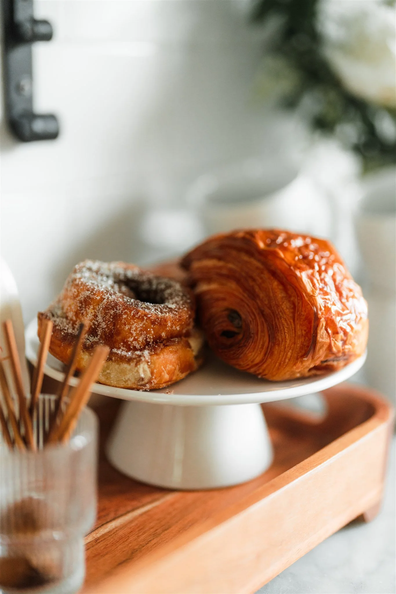 A white cake stand displays a glazed croissant and a cinnamon roll with sugar on top. A small glass jar with stir sticks is in the foreground, and a wooden tray is underneath the cake stand.