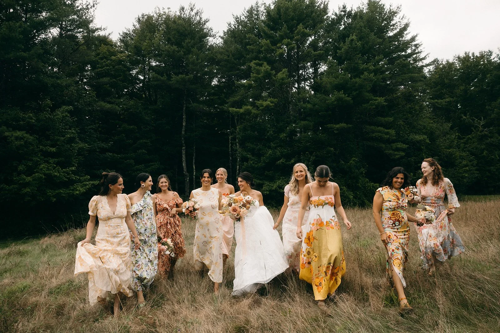 A group of women in floral dresses walking through a grassy field with trees in the background. One woman in a white wedding dress is at the center holding a bouquet. The women are smiling and appear to be celebrating.