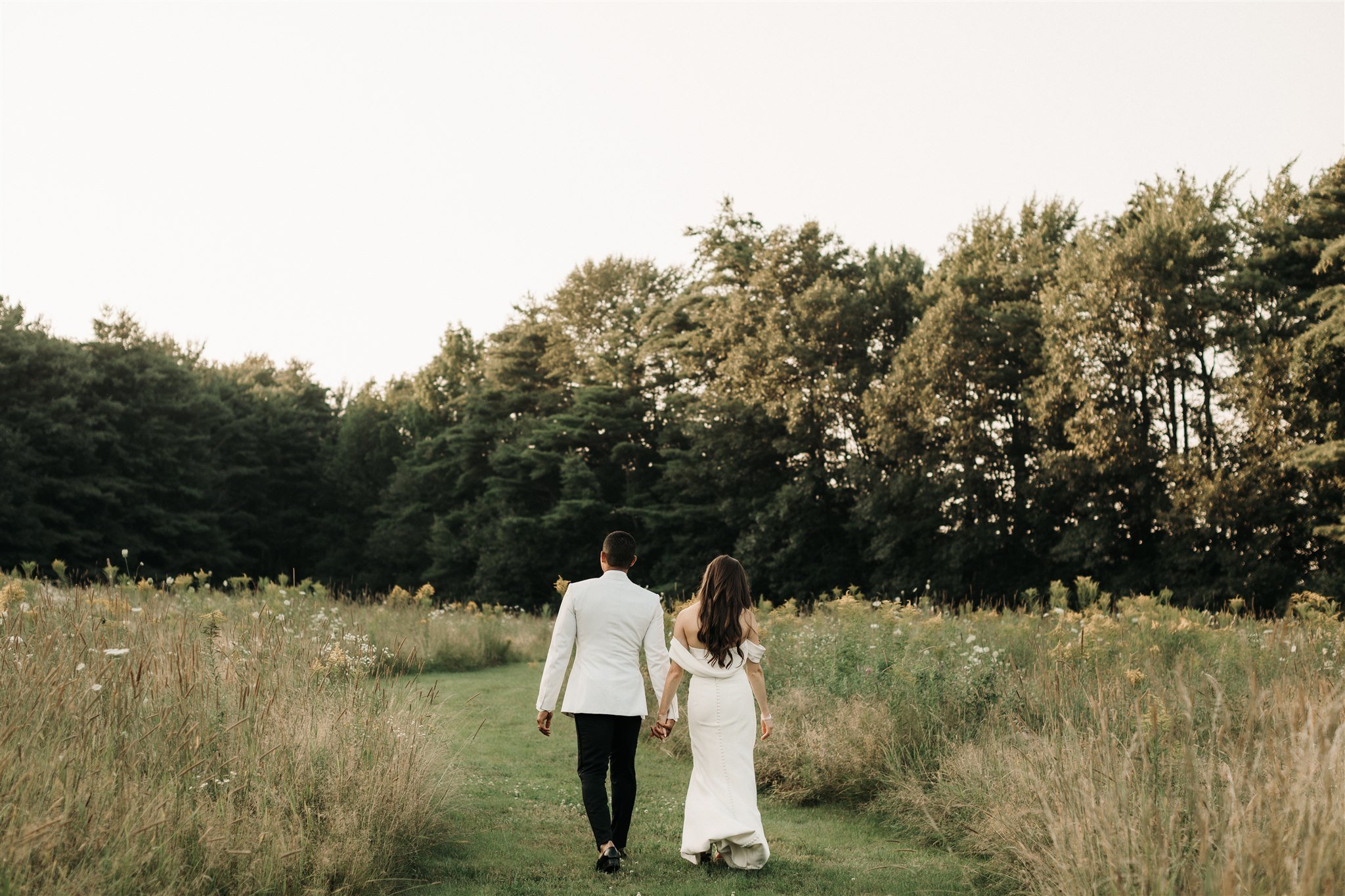 A couple in wedding attire walking hand in hand through a grassy field with trees in the background during sunset.