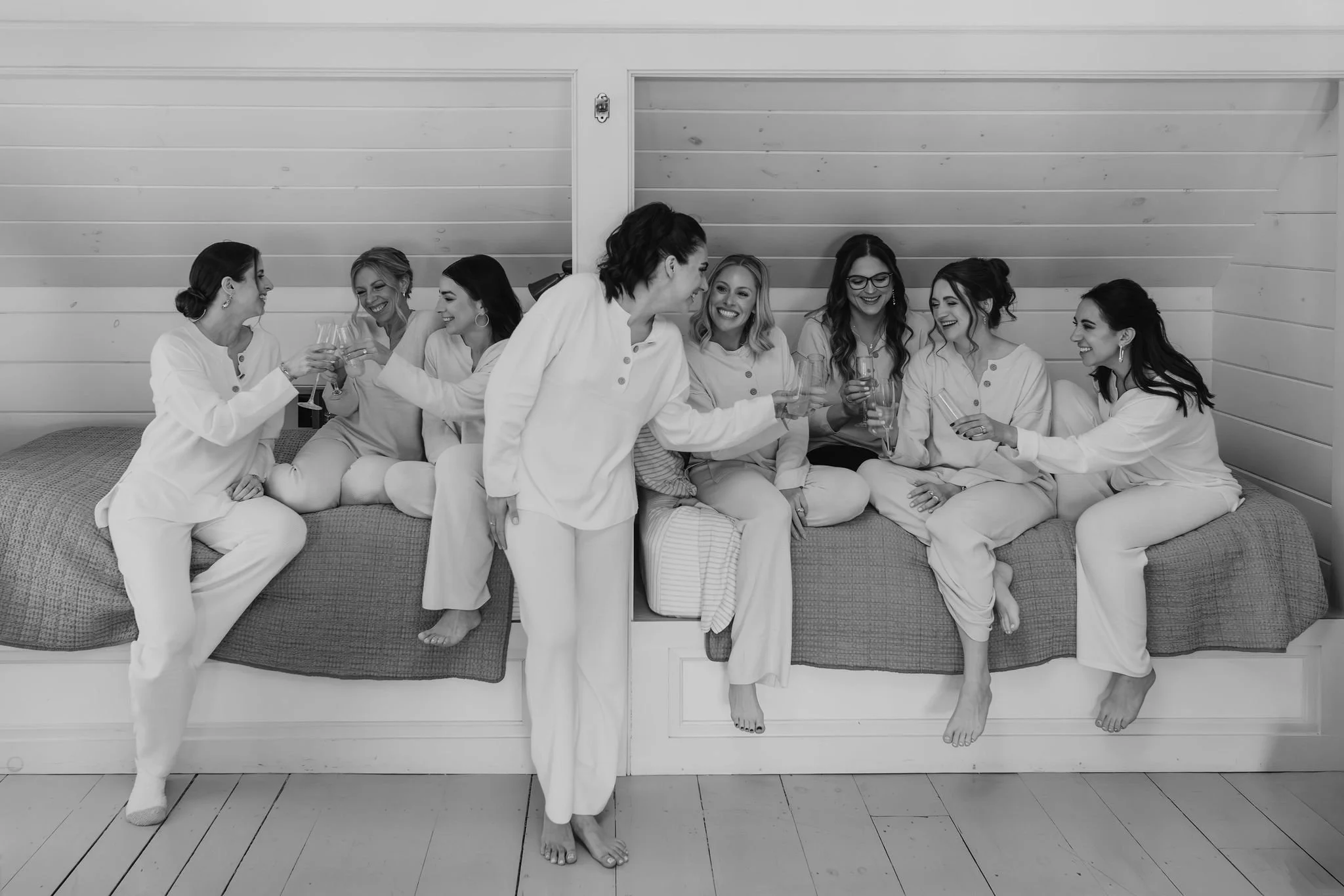 A group of eight women wearing matching pajamas, sitting and standing on a bed, smiling and toasting glasses in a cozy room with a wood-paneled ceiling.