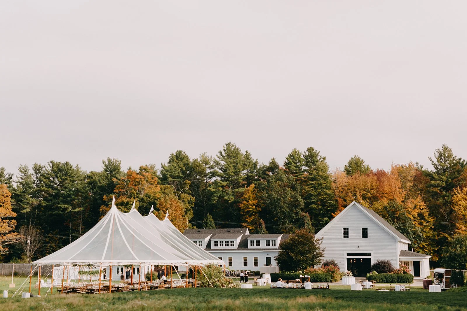 A large white event tent set up on a grassy lawn with wooden columns and drapes, surrounded by tables and chairs, near a white house with a barn-style door, with trees showing fall foliage in the background.