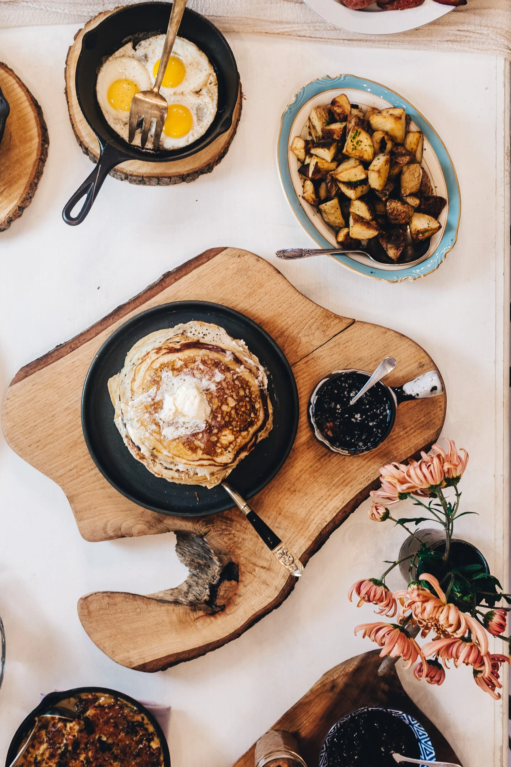 A breakfast table with a stack of pancakes with syrup and butter on a black plate, a small mug of syrup, a skillet of sunny side up eggs, roasted potatoes on a decorative plate, and pink flowers in a glass vase.