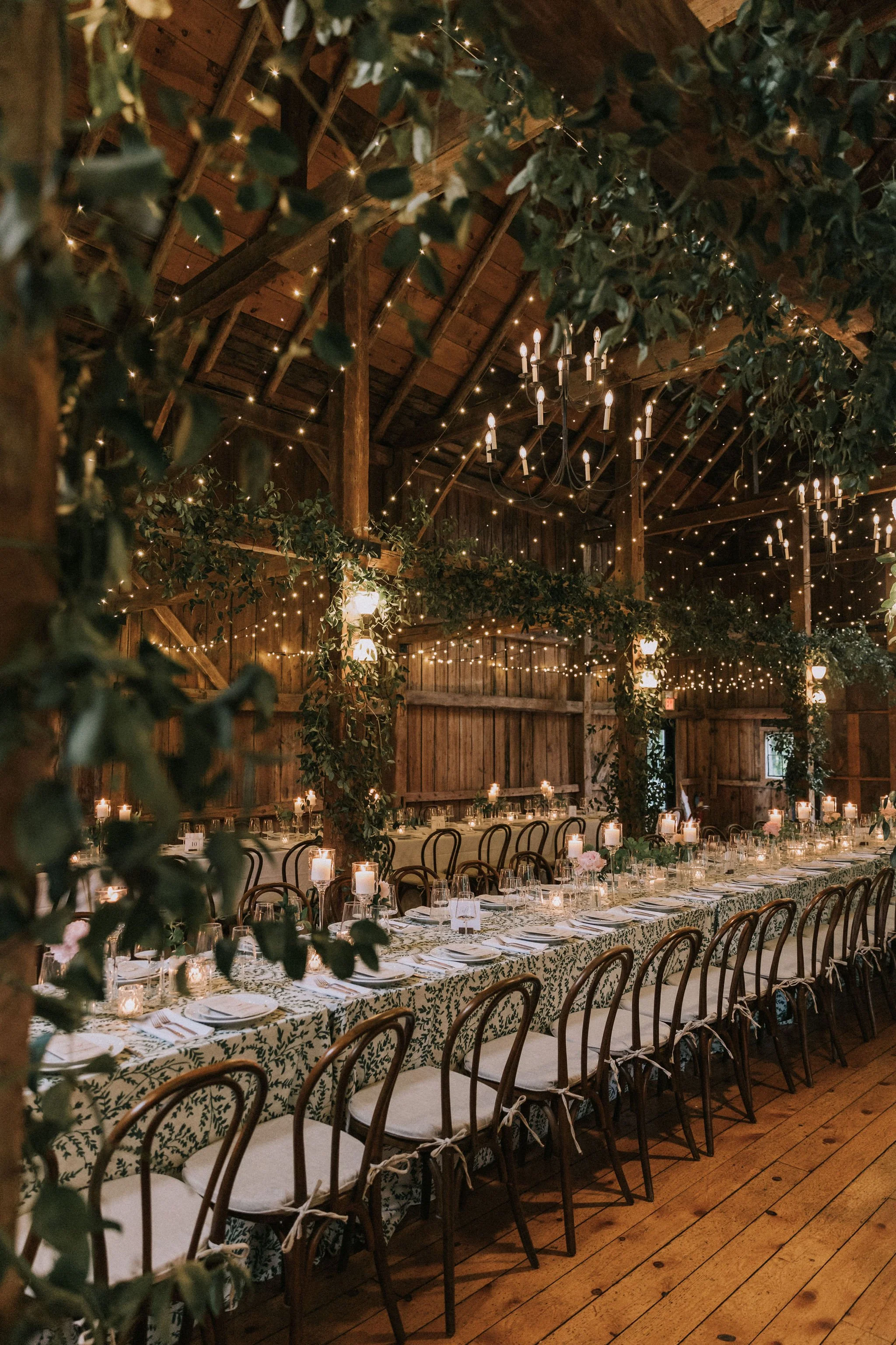 An elegantly decorated rustic banquet hall with a long table, candles, and string lights hanging from wooden beams.