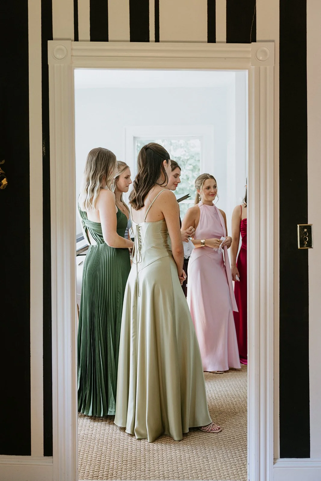 A group of women dressed in elegant gowns standing in a room, seen through a doorway. The women are in various shades of pink, red, green, and champagne, and are preparing for an event.
