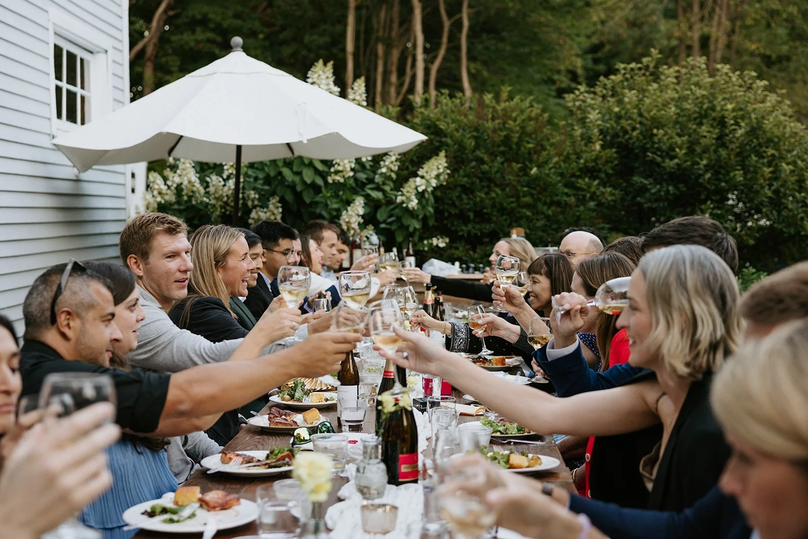 People sitting at a long outdoor dining table raising glasses in a toast during a celebration or gathering, with food and drinks on the table, umbrella overhead, and greenery in the background.