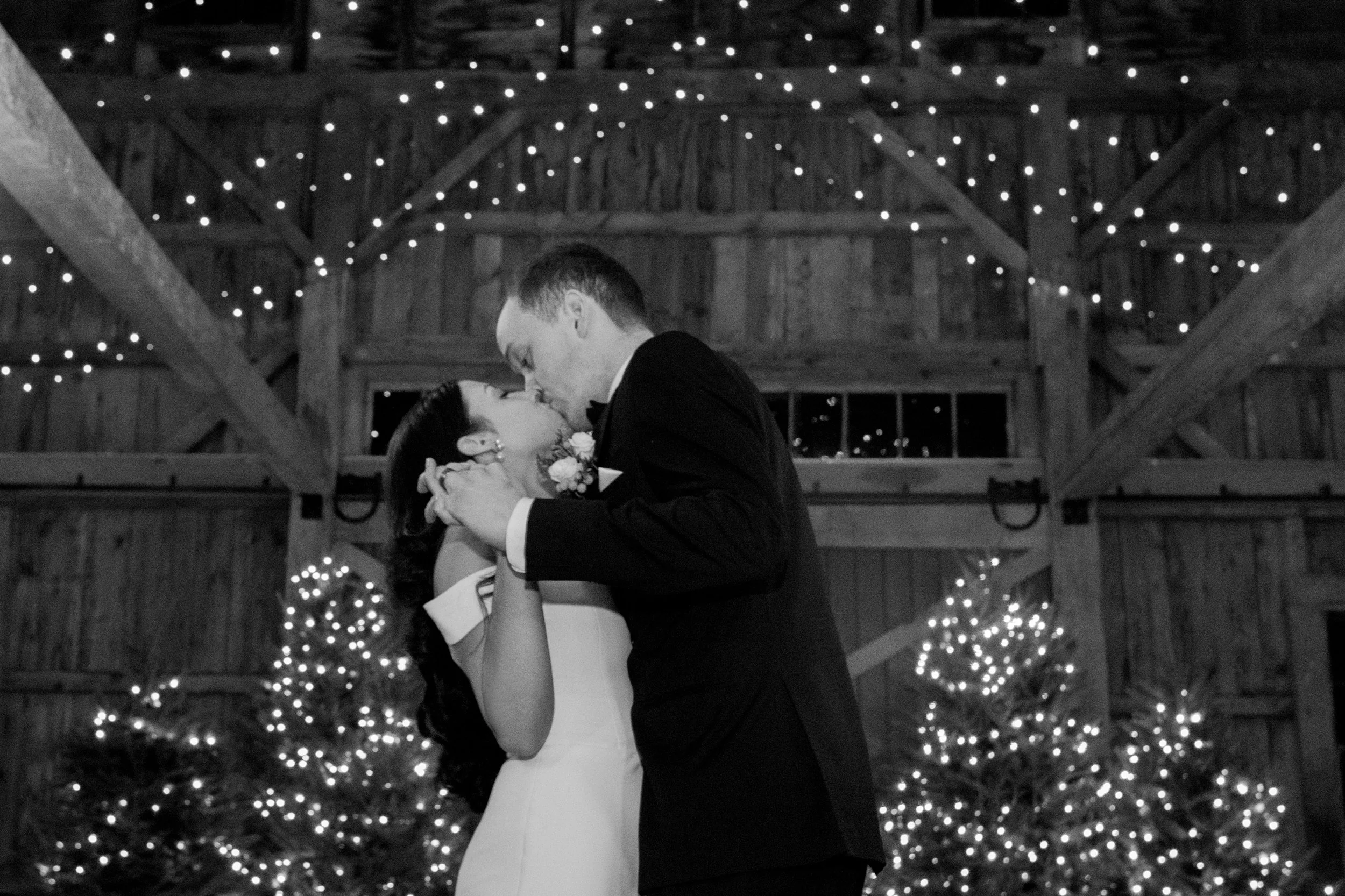 A black and white photo of a bride and groom sharing a kiss, holding each other closely inside a rustic barn decorated with string lights and Christmas trees.