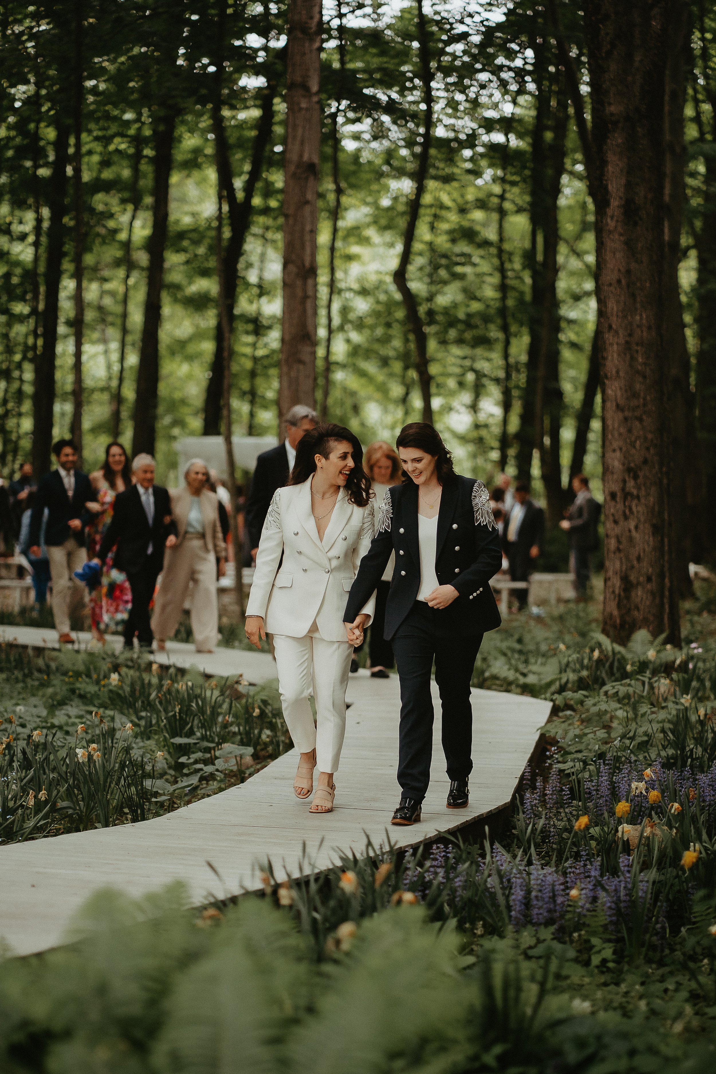 Two women holding hands and walking along a wooden pathway through a forest, surrounded by trees and flowers, with a group of people in the background.