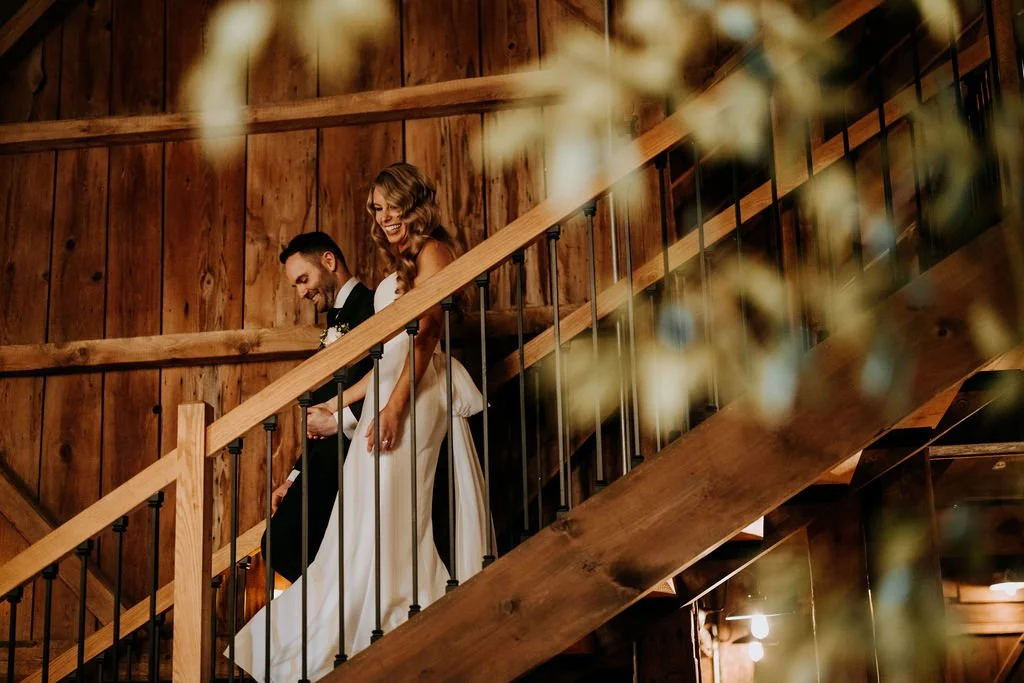 A bride and groom walking down a wooden staircase at a rustic wedding venue, smiling and holding hands.