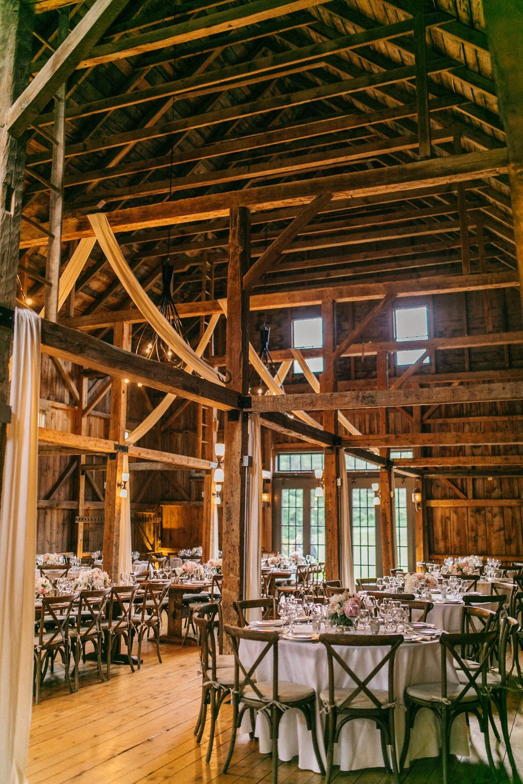 Interior of a rustic barn decorated for a wedding reception with round tables covered in white tablecloths, floral centerpieces, and wooden chairs. The high ceiling features exposed wooden beams and small windows, with soft warm lighting and draped f