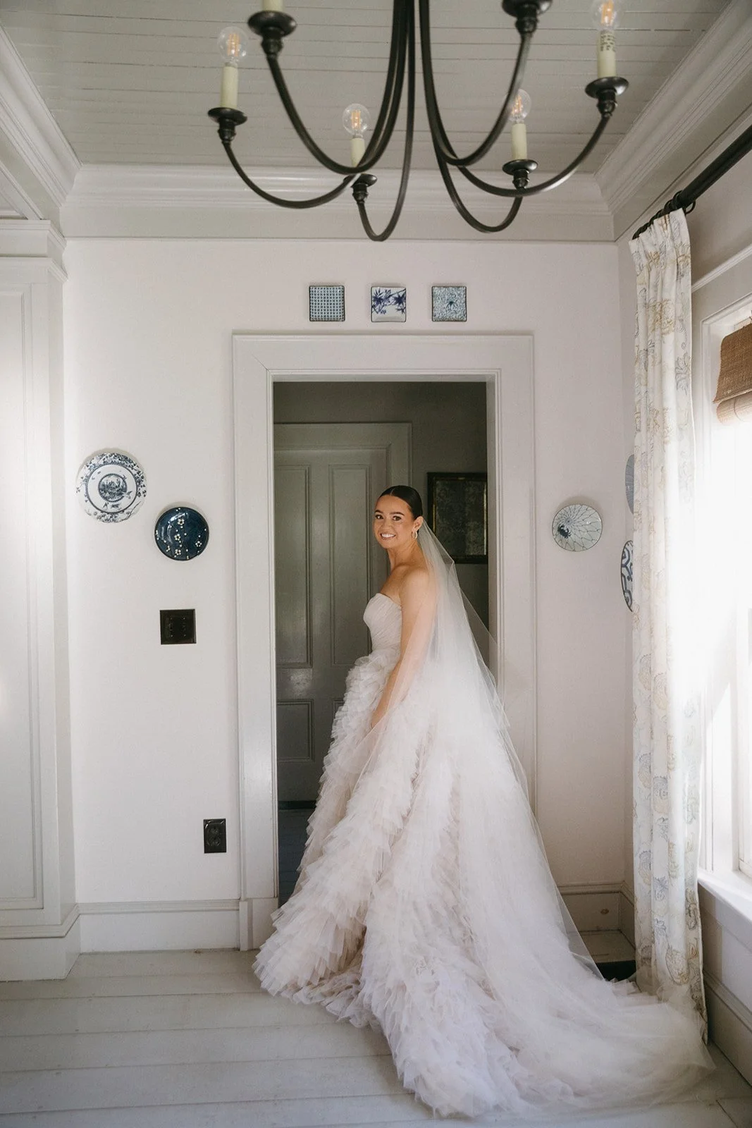 A smiling bride in a strapless white wedding dress with a long, flowing tulle skirt stands in a doorway in a bright room with white walls and vintage decor, including plates and small framed art, with a chandelier hanging from the ceiling.