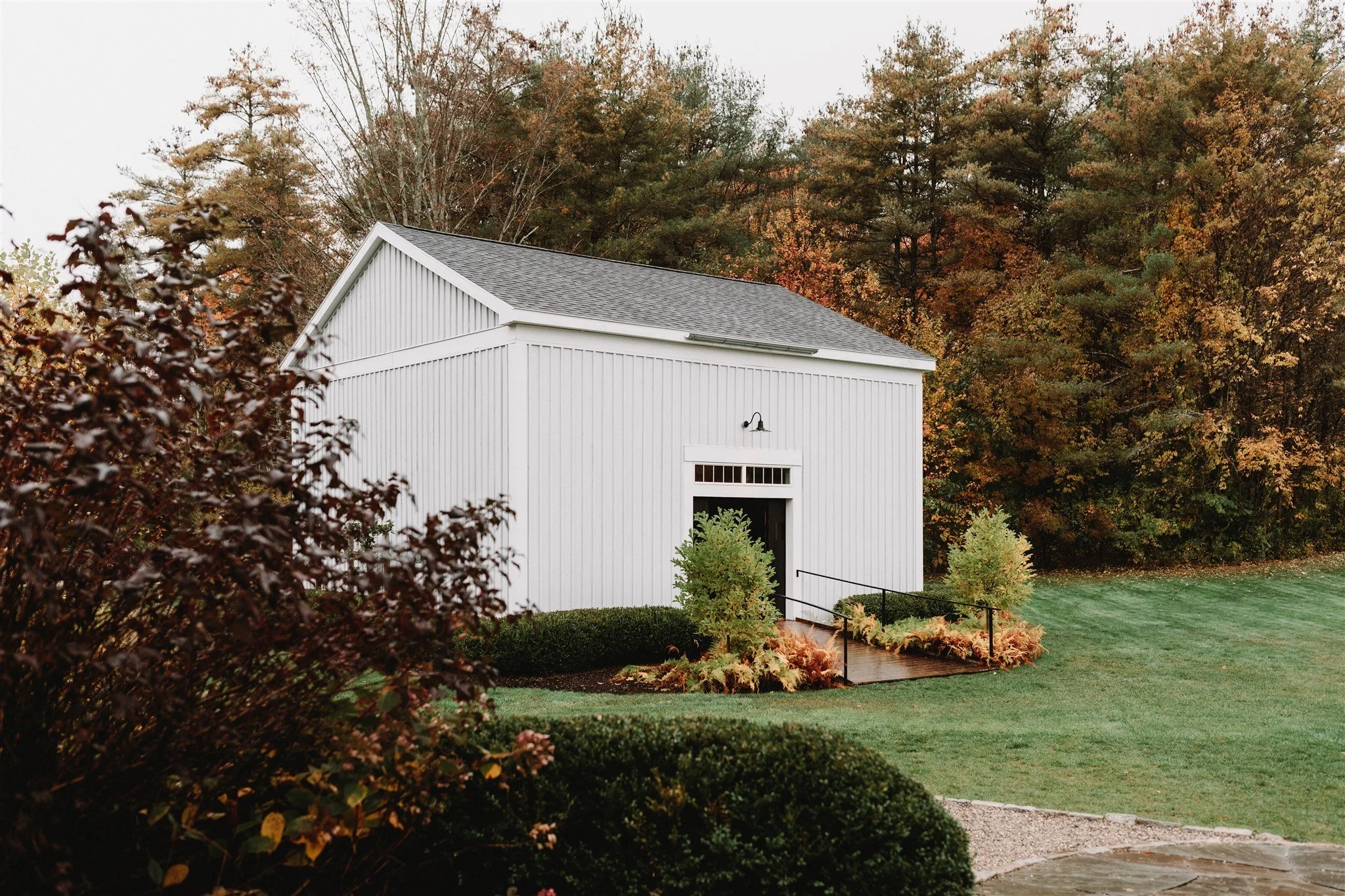 A white barn with a small window near the roof and a black door, surrounded by green trees with autumn foliage, and a landscaped area with bushes, plants, and a small wooden ramp.