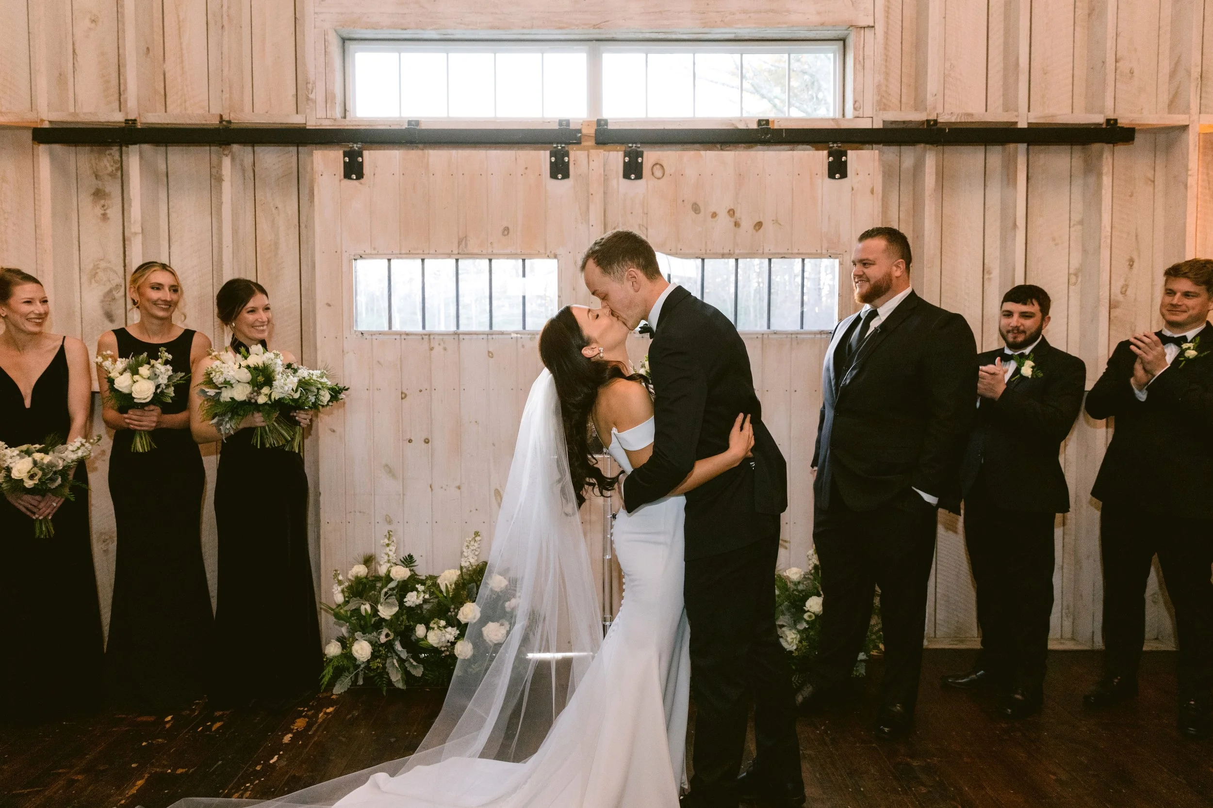 A wedding ceremony with a bride and groom sharing a kiss, surrounded by bridesmaids and groomsmen clapping in a rustic barn setting.