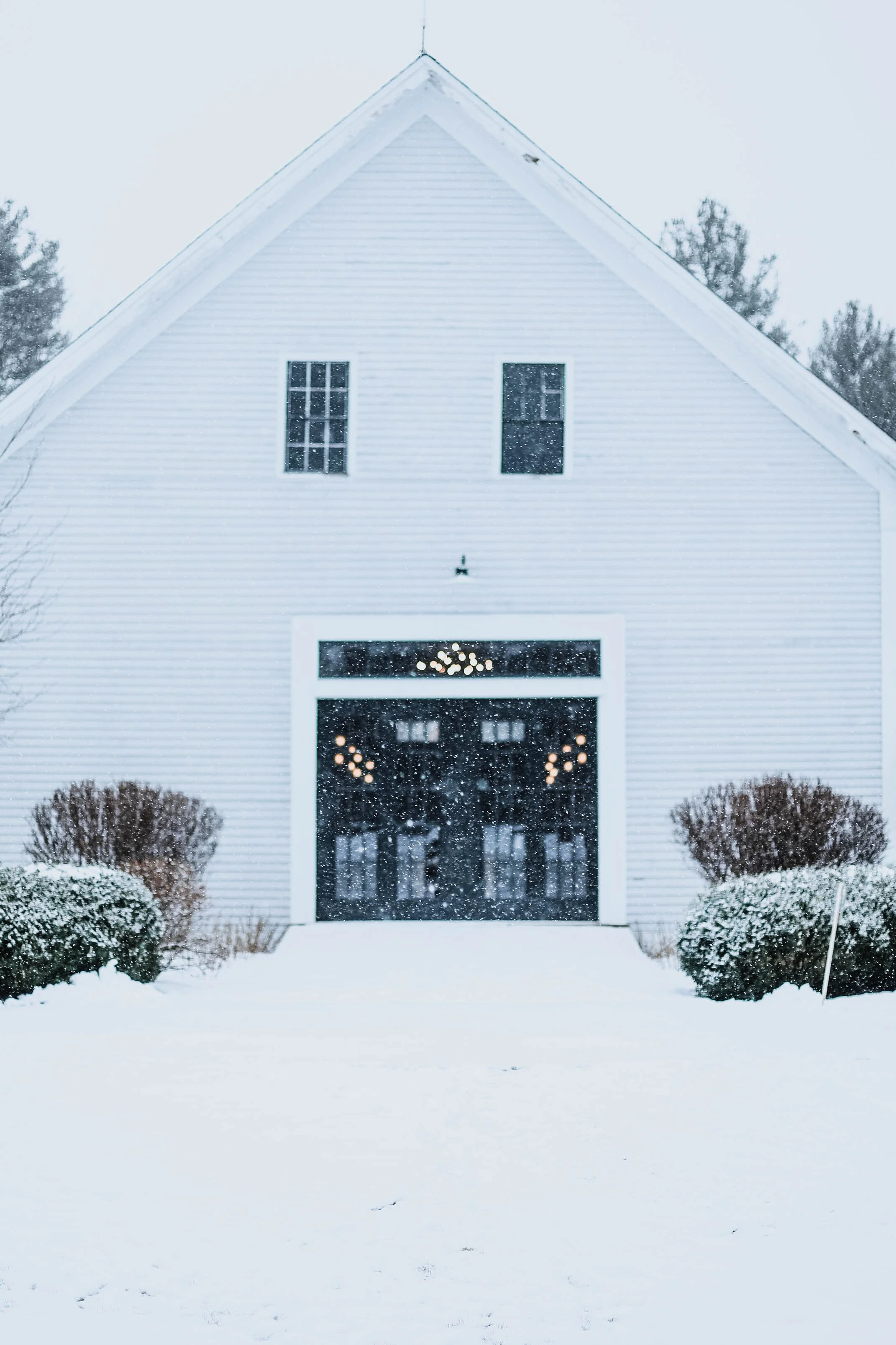 White barn with hanging string lights and snow falling outside.