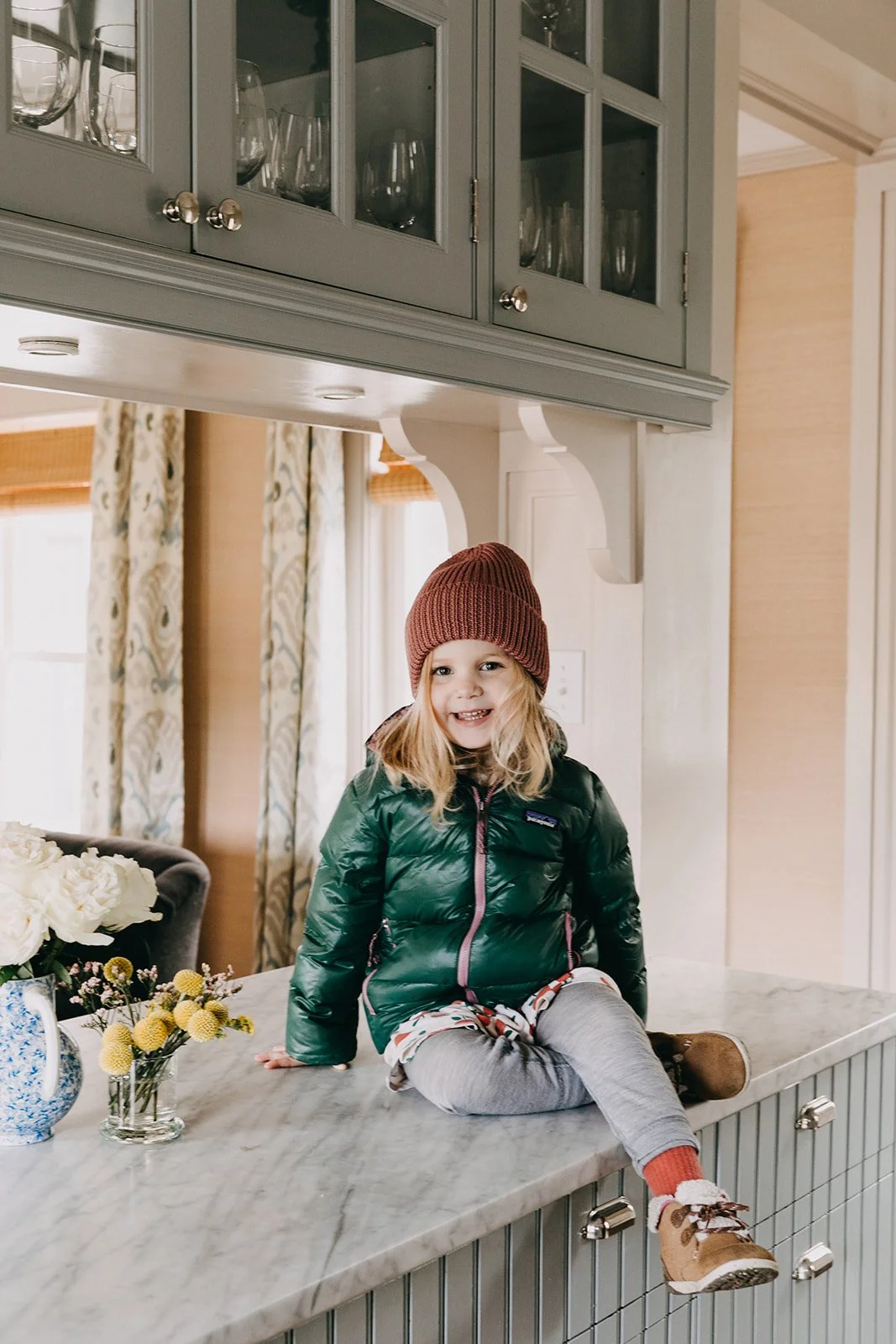 A young girl with blonde hair, wearing a red beanie, green puffer jacket, gray leggings, and tan boots, sitting on a white marble kitchen counter with a bouquet of white and yellow flowers nearby. She is smiling at the camera in a cozy, well-lit kitc