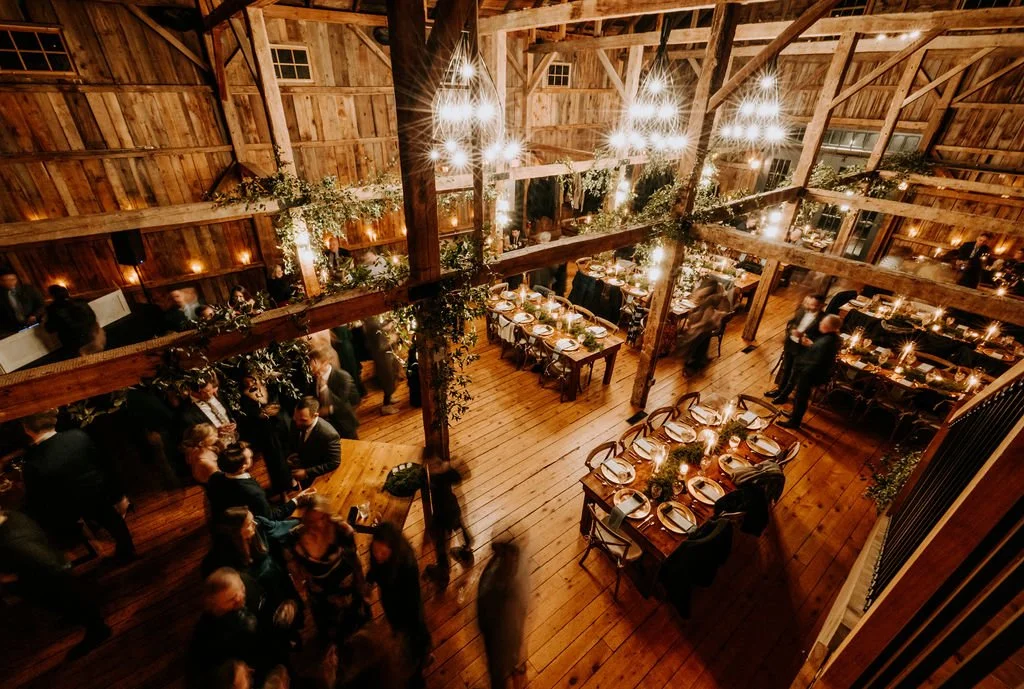 A cozy rustic wooden barn decorated with string lights, long tables set for a dinner party, and people socializing at an evening event.